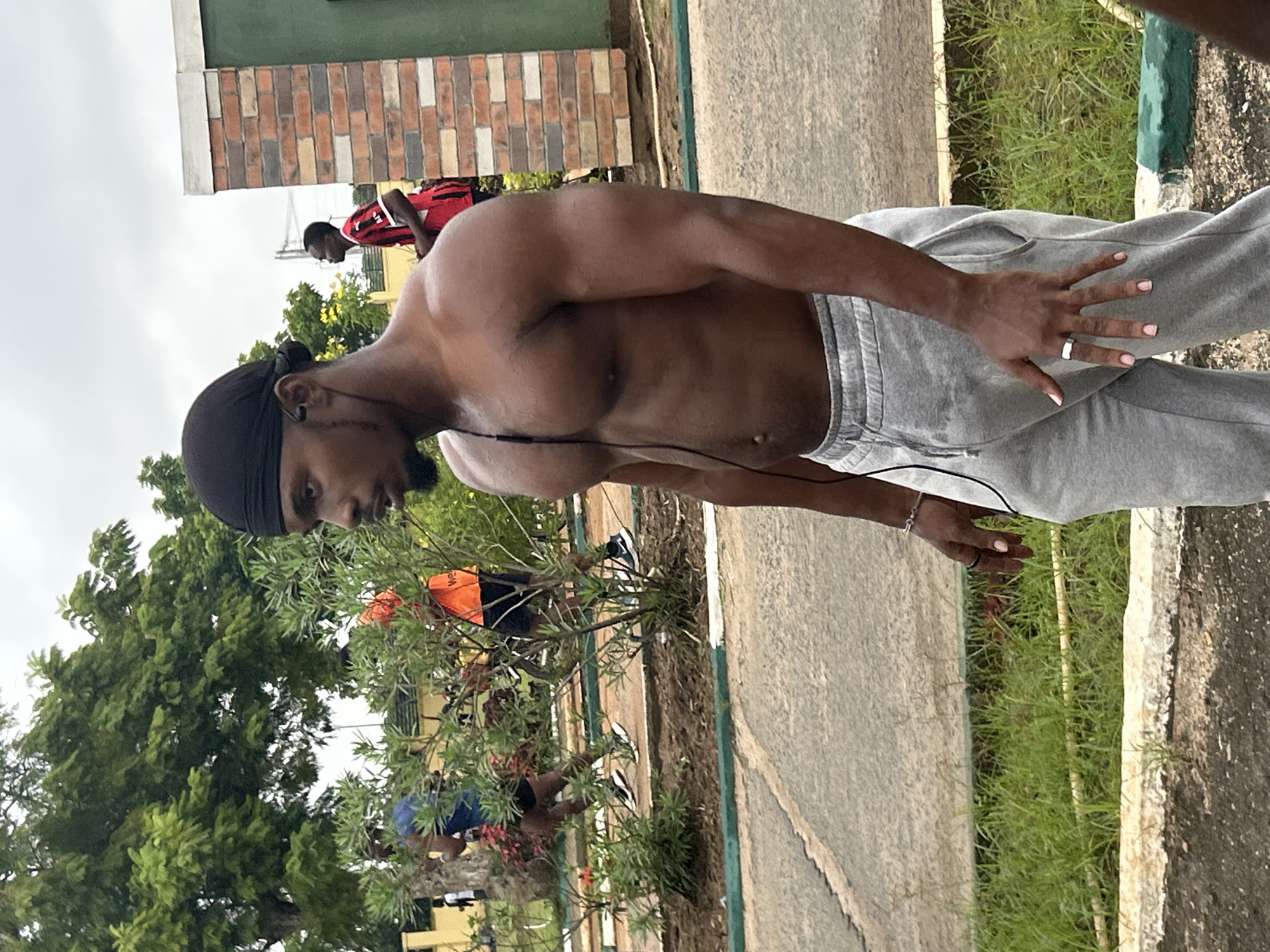 The image depicts a shirtless Black man wearing a black durag and grey sweatpants, with earbuds in his ears, standing on a paved area with green grass and a dirt path in the background. He is facing right, with his left arm bent at the elbow and his left hand held up, revealing a silver ring on his ring finger. His right arm is down by his side. The man's skin is dark brown and appears sweaty, suggesting physical exertion. In the background, other individuals are visible, some appearing to be running or exercising. One person in the middle ground wears an orange vest, and another in the further background wears a red and black striped shirt. A brick building with a green accent is partially visible on the right side of the frame. The overall setting appears to be an outdoor recreational or athletic facility. The presence of lush green trees and vegetation suggests a tropical or subtropical climate, consistent with the location of Ihiagwa, Nigeria. The sky is overcast with a light grey color. The ground is a mixture of asphalt and concrete with visible grass and some scattered debris.