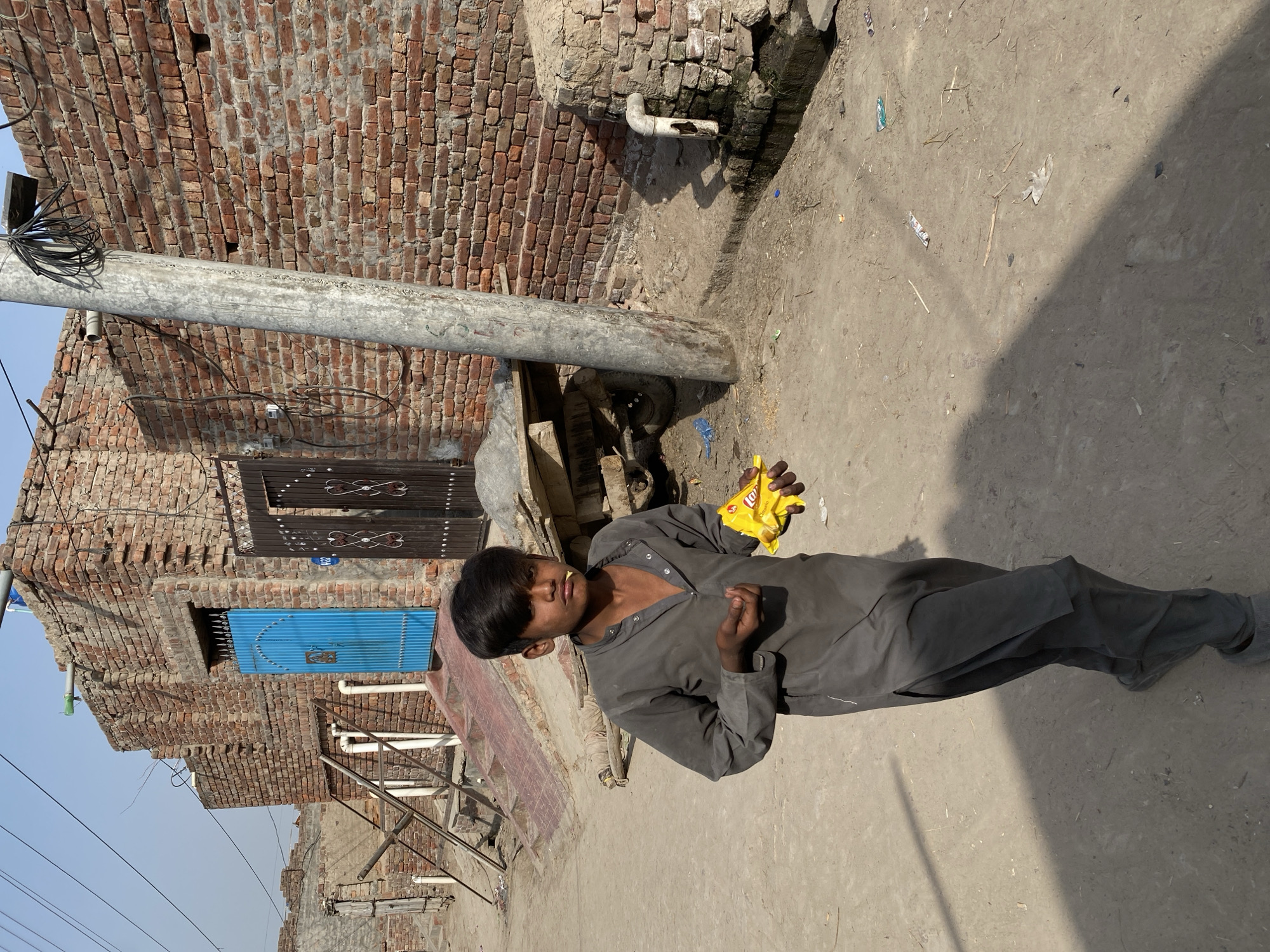A young boy stands in a dusty alleyway or street, likely in Chak 166 GB, Pakistan, under bright daylight. He wears a plain grey collared shirt and matching trousers. In his left hand, he holds a bright yellow bag of Lay's potato chips, with his right hand near his mouth, suggesting he is eating or about to eat.
The background consists of buildings made from reddish-brown bricks, with a general appearance of wear. A tall, grey metal pole with numerous bundled electrical wires stands prominently on the left. Two doors are visible on the brick structure: a vibrant blue corrugated metal door on the lower level and a dark wooden door with ornate patterns and the number "242" inscribed on it, higher up.
The ground is unpaved, covered in dry, light-colored earth with some scattered small debris. Distinct shadows are cast, indicating a clear, sunny day. The boy's expression is neutral as he looks towards the camera. Visible text includes "Lay's" and "Potato Chips" on the yellow snack bag and the number "242" on the dark wooden door.