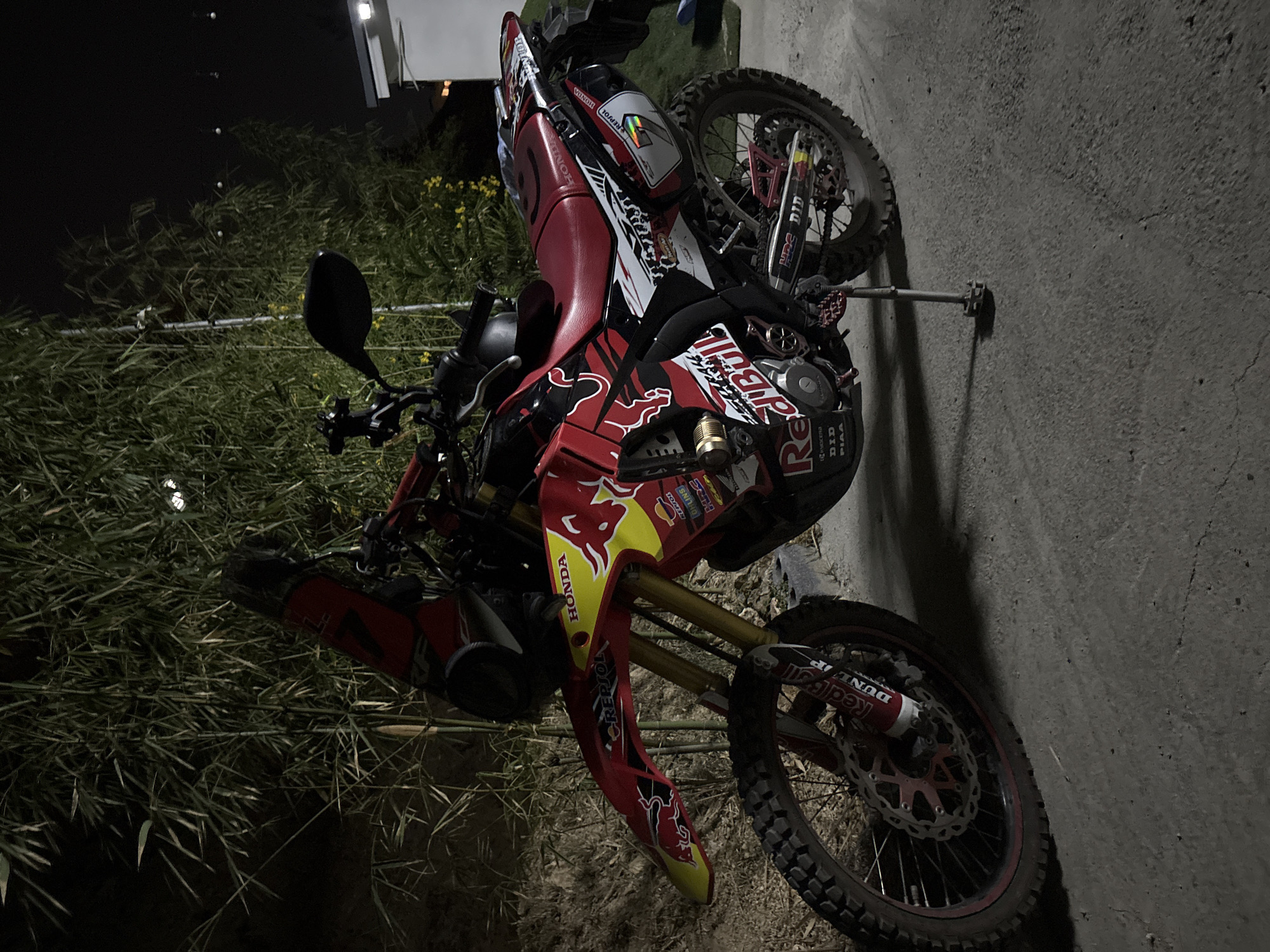 The image depicts a Red Bull-branded Honda dirt bike resting on its kickstand on a dark paved surface, possibly a road or parking area, in what seems to be an outdoor setting. The background includes tall grass or reeds. The text "HONDA," "REPSOL," "Red Bull," "KYOCERA," "DID PIAA", "OHLINS" and "DUNL IP" are visible on the bike's body. Although a specific location such as Baan Bang Rak, Thailand is mentioned, there is no visual information in the image to verify this detail. The scene is stationary and the bike is the central subject of the image.