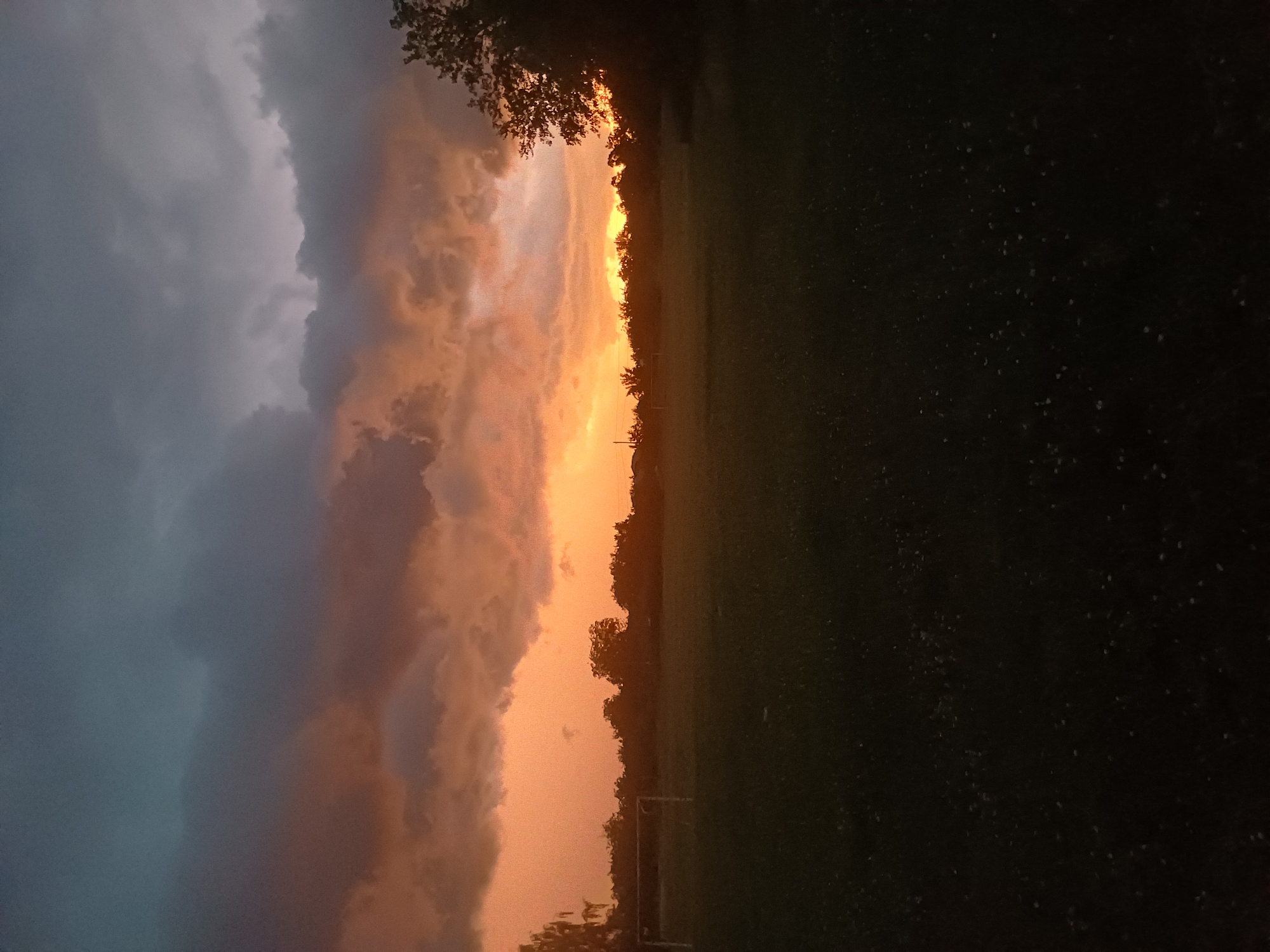 This image captures a dramatic sky over a field, likely at sunset or sunrise, given the intense orange and pink hues illuminating the clouds. The sky on the left is dominated by dark, brooding clouds that transition into a vibrant band of sunset colors towards the horizon. Silhouetted trees form a dark line along the edge of the field, with hints of leaves visible against the bright sky.
The field itself appears to be a grassy area, possibly a park or sports field, with a soccer goal visible in the mid-ground. The grass is dark, suggesting it is either evening or the lighting conditions are low. A few scattered lights or stars are visible in the dark areas of the image, potentially indicating the presence of fireflies or distant lights.
The overall mood is one of natural beauty and awe, with the juxtaposition of the dark, stormy clouds and the radiant sunset. The scene evokes a sense of the transition from day to night, with a powerful display of atmospheric phenomena. The location is identified as Trypillia, Ukraine, which adds a geographical context to the otherwise universal depiction of a striking sky. There is no visible text in the image.