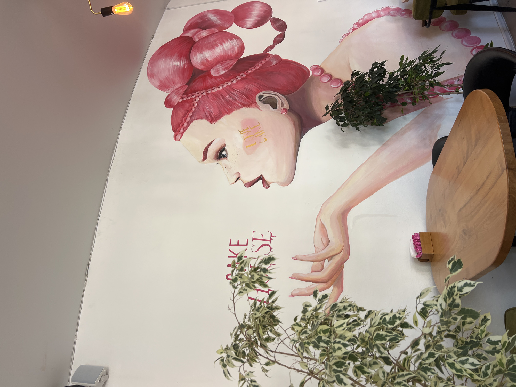 The image depicts the interior of a cafe in Odesa, Ukraine, featuring a vibrant mural on a white wall. The mural is an artistic representation of a woman with bright pink hair styled in elaborate buns and braids, adorned with a pearl necklace. She is shown in profile, with her mouth open as if blowing a kiss or exhaling. The words "CAKE HOUSE" are painted in pink on the wall, with dripping effects, and the words "LOVE CAKE" are visible on the woman's cheek in gold lettering.
In the foreground, a light-colored wooden table occupies the lower half of the frame. On the table, a small wooden holder contains packets of pink sugar or sweetener. To the left of the table, a lush green plant with variegated leaves, possibly a ficus, is positioned. Another smaller, dark green plant is placed near the right edge of the table, partially obscuring the mural. The cafe appears to have a modern and artistic ambiance, with a prominent light fixture with a yellow bulb hanging from the ceiling in the upper right corner. A white speaker is visible mounted on the wall in the upper left corner. The overall scene suggests a cheerful and inviting atmosphere.
