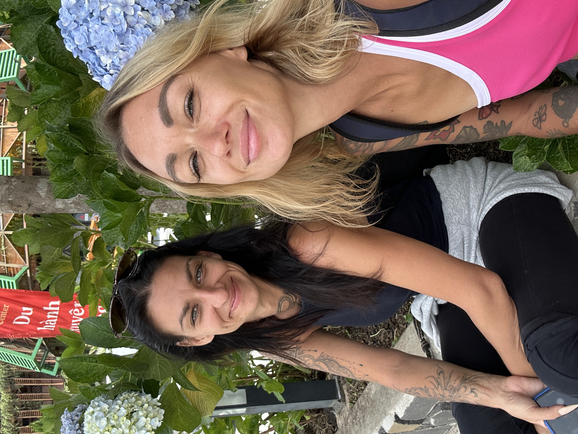 Two women are seated outdoors in front of lush green foliage and blooming hydrangeas. A red banner with Vietnamese text, "Center Du hành xuyên...", is visible in the background. The woman on the left has dark hair and is wearing a black tank top and dark leggings. She has tattoos on her arm and neck and is holding a mobile phone. The woman on the right has blonde hair and is wearing a pink and white athletic top with a dark backpack. Both women are smiling at the camera. The scene suggests a relaxed, outdoor setting, possibly a garden or park in Da Lat, Vietnam, known for its hydrangeas and scenic landscapes.