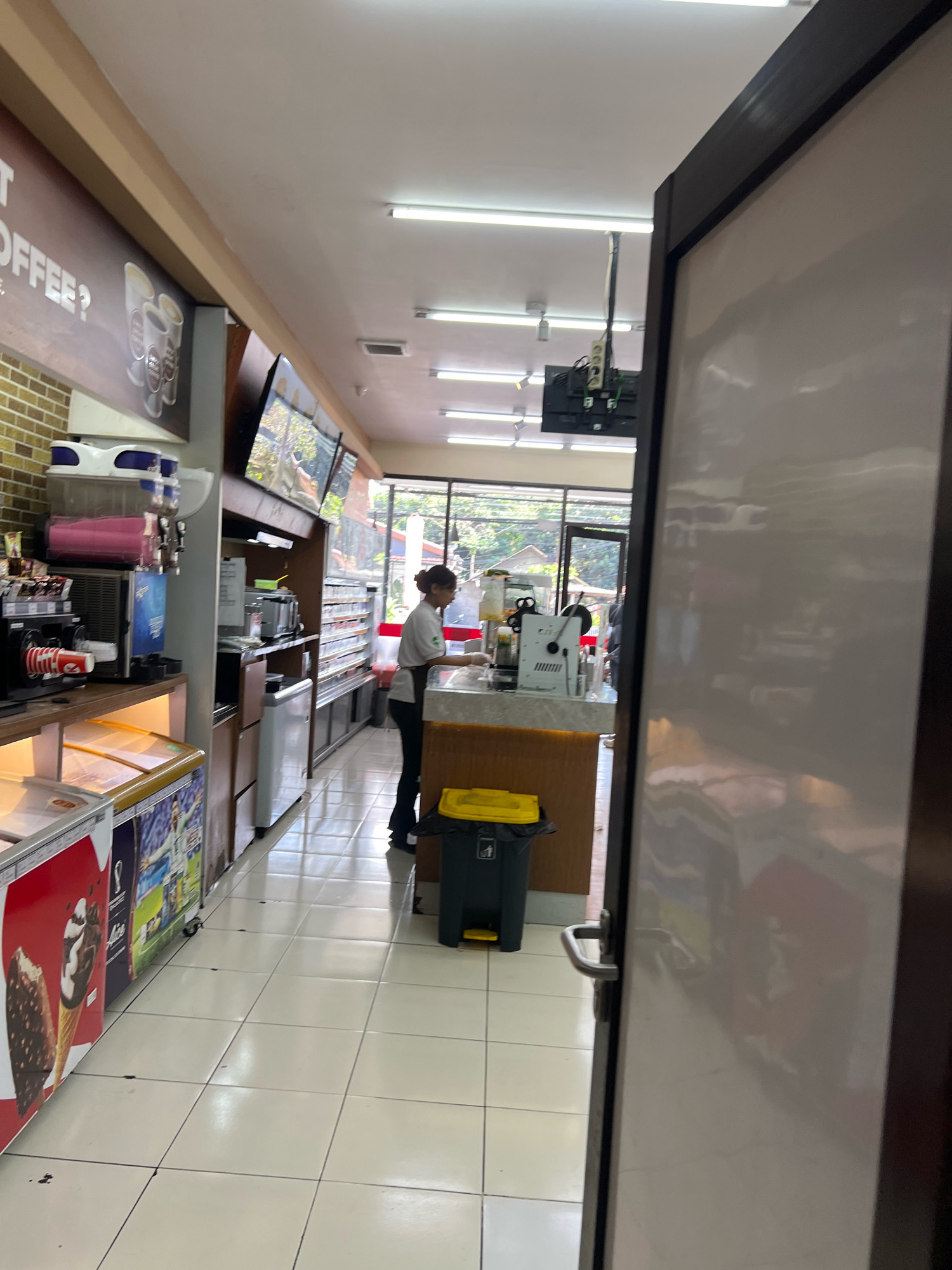 The image captures the interior of a convenience store, likely a 7-Eleven, in Denpasar, Indonesia. The perspective is from just inside the entrance, looking down a narrow aisle. On the left, there are refrigerators displaying ice cream and beverages, adorned with colorful advertisements. Further down, a counter area is visible with a cashier wearing a white shirt and apron, attending to a customer or preparing an order. Behind the counter, shelves are stocked with various products, and a digital screen displays advertisements. The floor is tiled in a checkered pattern. The overall atmosphere suggests a typical urban convenience store setting.