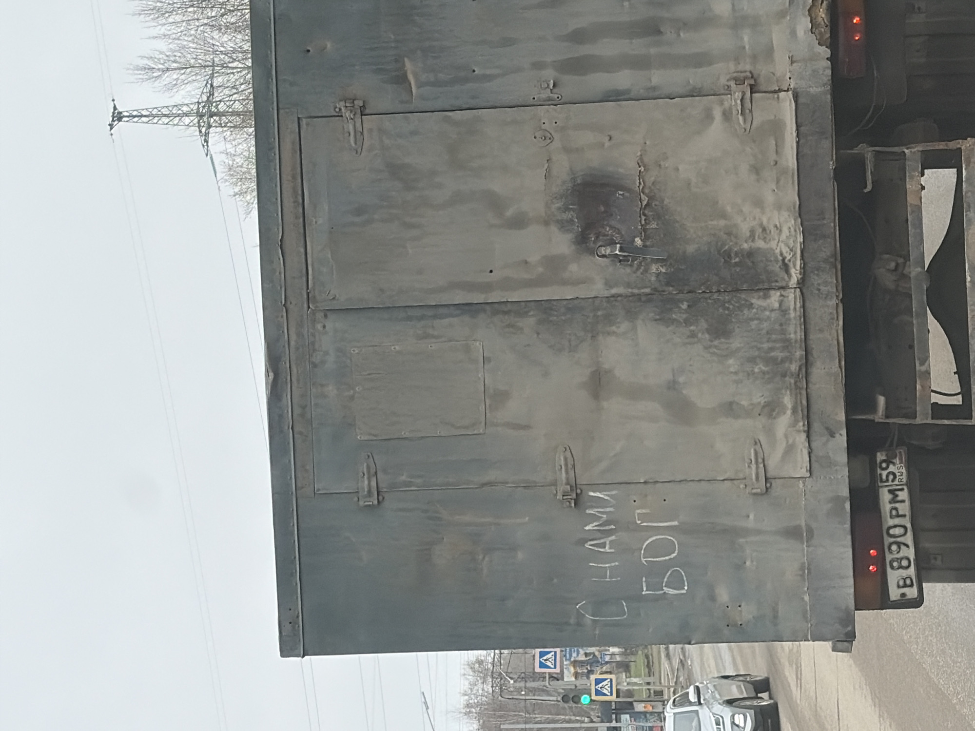 The image shows the rear of a dirty, metallic-gray truck with a rectangular cargo box, taken in Chaikovsky, Russia. The truck's license plate is visible, reading "B 890 PM 59 RUS." The cargo box is divided into sections with latches. Scrawled graffiti in white lettering on the lower right side of the cargo box reads "C HAMA БОГ." The background features a cityscape including pedestrian crossing signs, a traffic light showing a green light, and other vehicles. Power lines and bare trees are also visible.