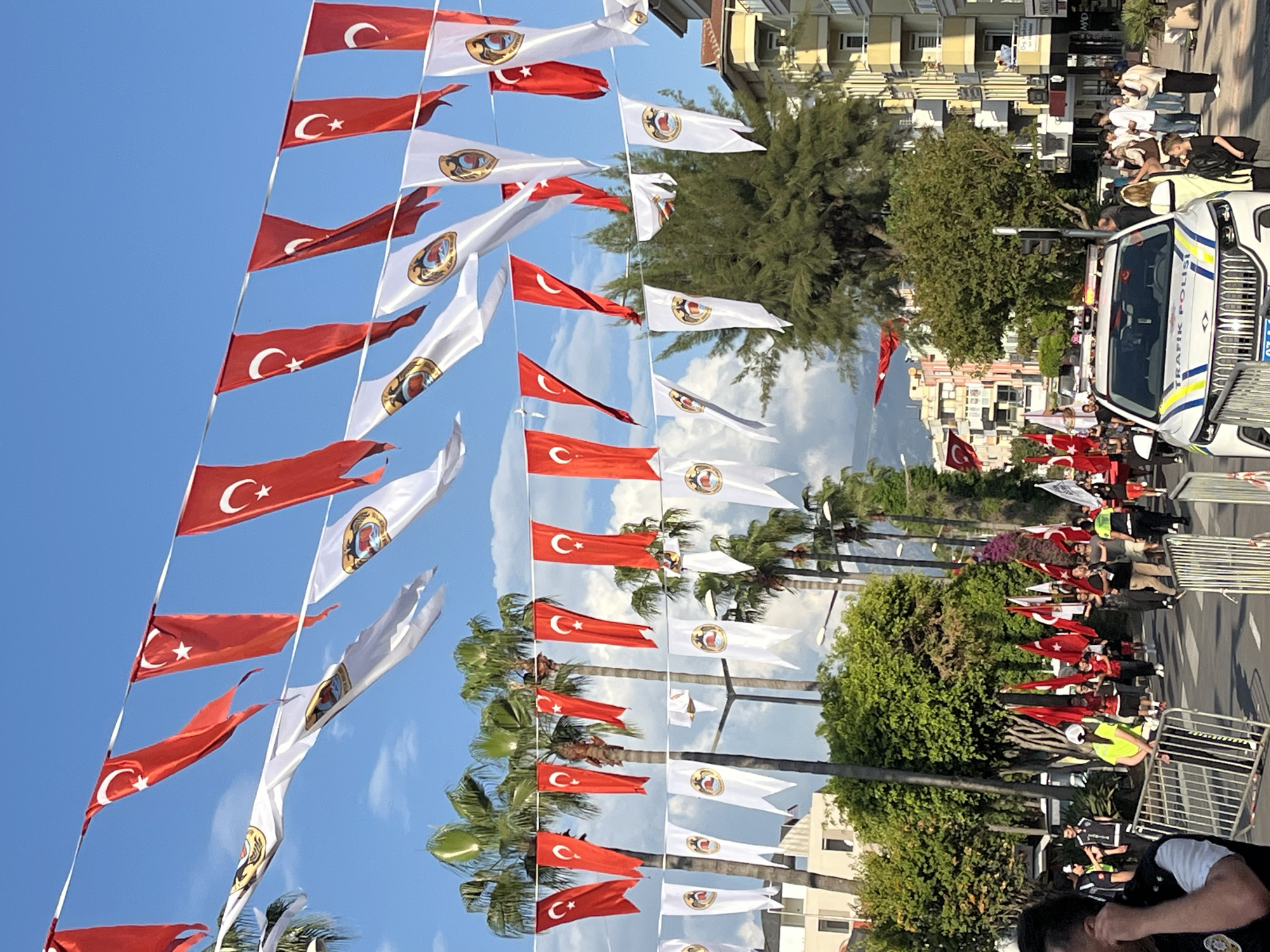 The image shows a street in Alanya, Turkey, decorated with Turkish flags and white flags bearing a circular emblem. A police car is parked on the right side of the street. People are standing along the street behind metal barriers, some holding flags. Palm trees line the street, and buildings are visible in the background. The sky is blue.