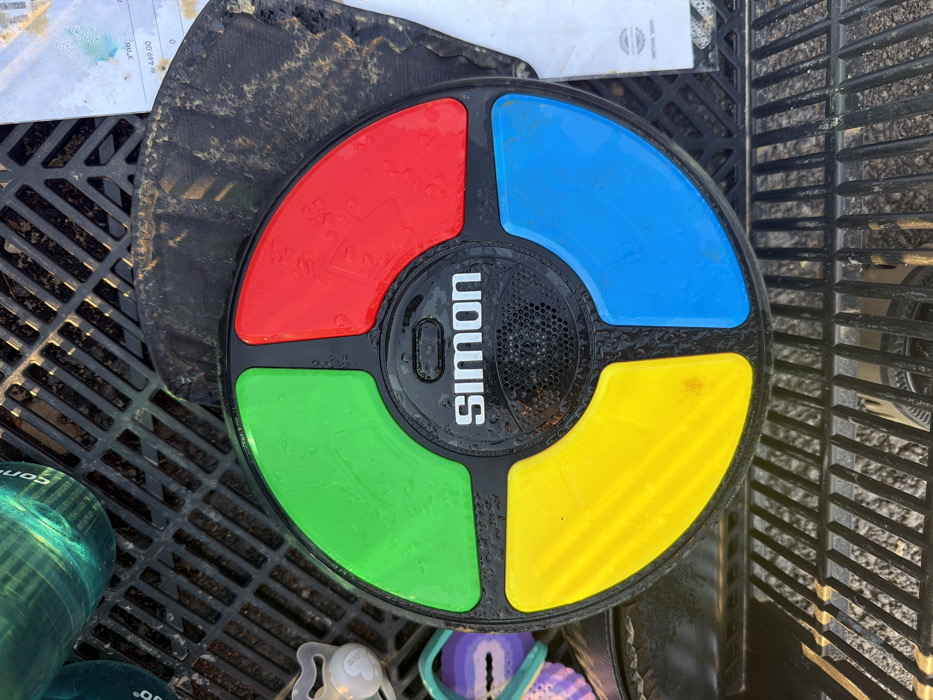 A Simon electronic memory game sits on a black plastic grid. The game is round with a black base and four colored quadrants: red, blue, green, and yellow. The word "SIMON" is prominently displayed in white letters above a speaker grill in the center. Droplets of water are visible on the surface. The background includes other objects such as a dirty black square, a piece of paper, and a plastic container.