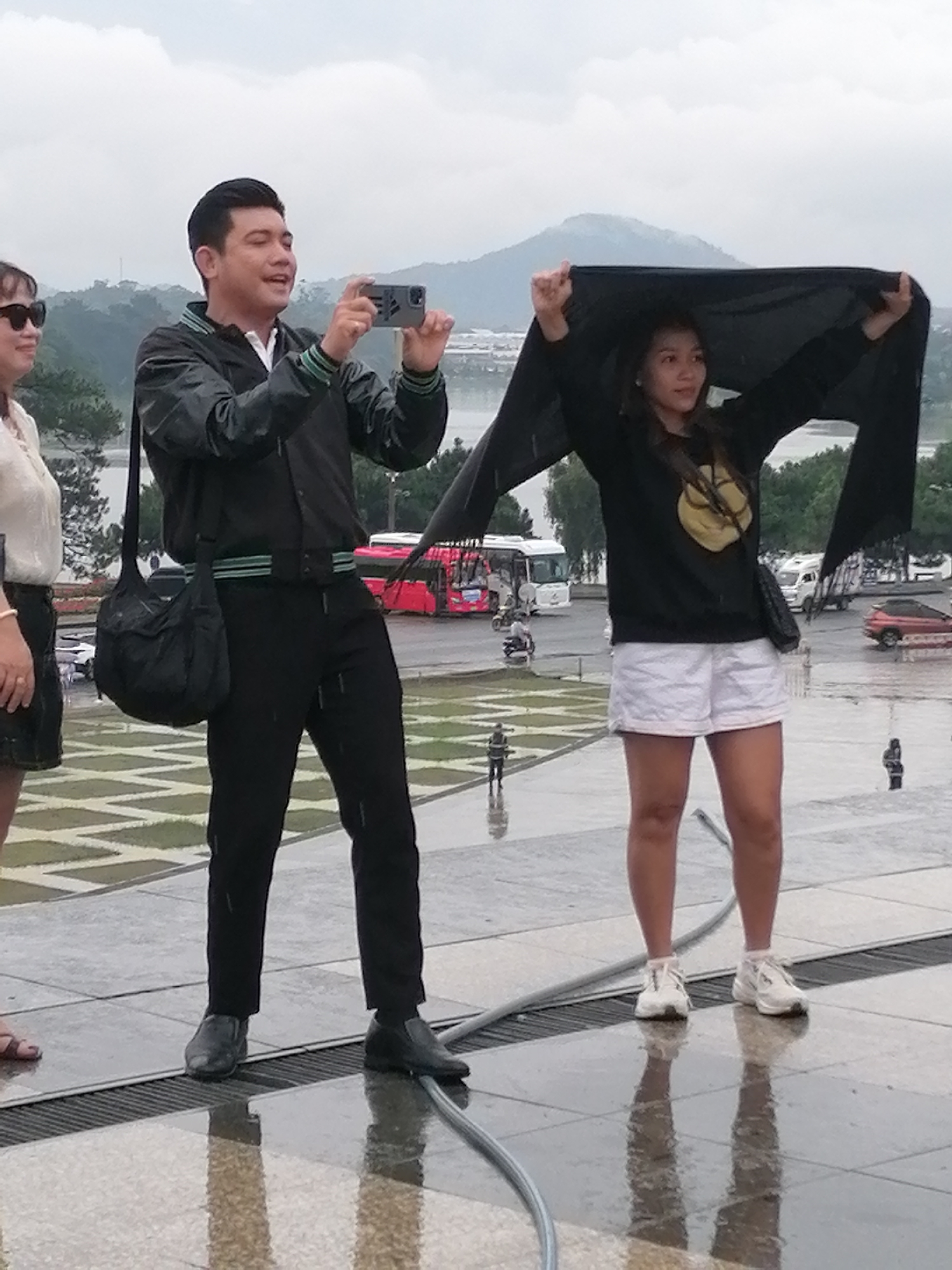 A man and a woman stand on a wet, patterned surface, likely outdoors in Da Lat, Vietnam, given the overcast sky and lush green hills in the background. The man, on the left, is wearing a black jacket with green and white trim, a white shirt, and black pants. He is holding a smartphone with both hands and appears to be taking a photo. A black messenger bag hangs from his left shoulder. To his right, the woman is holding a large black piece of fabric over her head, creating a makeshift umbrella. She is wearing a black long-sleeved top with a circular yellow design on it, white shorts, and white sneakers. The ground is wet and reflective, suggesting recent rain. In the background, there are trees, a body of water, and several vehicles, including a red bus and a white van. The overall atmosphere is one of a cloudy, possibly rainy, day.