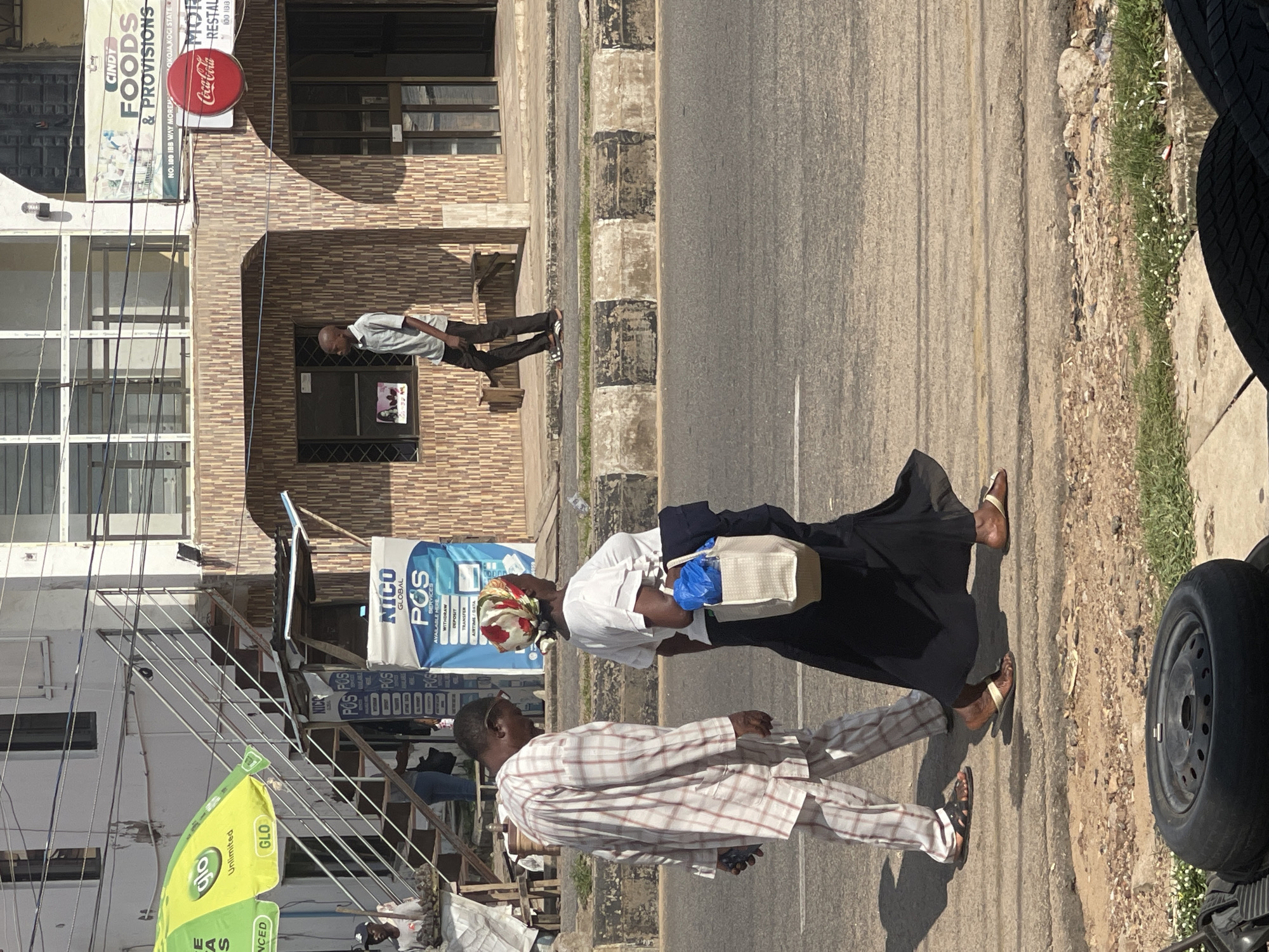 The image shows a street scene with several people and buildings. In the foreground, a person in a plaid suit is walking towards the left, while another person in a white shirt and black skirt walks away from the camera. They appear to be on a sidewalk next to a road. In the background, there are buildings with shops, including one with signs for "CINDY FOODS & PROVISIONS" and "Coca-Cola RESTAL". There's also a blue sign that says "NICO POS GLOBAL SERVICES". The lighting suggests it is daytime, possibly late afternoon due to the long shadows. The scene appears to be in an urban or semi-urban area, with a mix of commercial establishments and street activity.