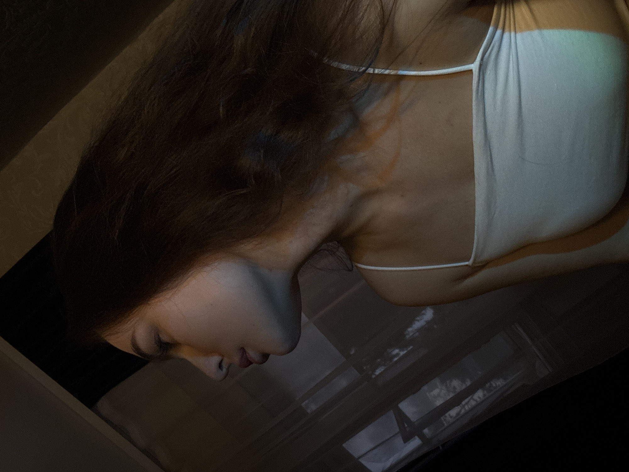 A side profile of a young woman with long, dark hair is visible, her face turned to the left. She is wearing a white, sleeveless tank top. The background appears to be indoors, with a dark, shadowed area on the left and a window with sheer curtains to the right, through which tree branches are faintly visible. There is a diagonal line of light and shadow across the woman's chest, suggesting indoor lighting or light coming from the window. The scene does not provide any specific information to suggest a location in Chaikovsky, Russia.