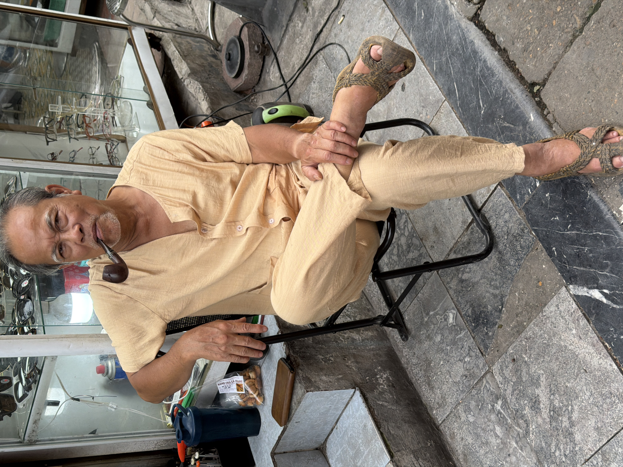 The image depicts an older Asian man sitting outdoors in what appears to be Hà Nội, Vietnam. He is wearing a light brown or beige outfit and sandals, and is smoking a pipe. He is seated on a small, foldable metal chair. His right foot is placed on his left thigh, and his left hand is resting on his right leg. He is sitting adjacent to a glass display case filled with eyeglasses. Behind him are various objects, including a grinding stone, and various power cables. The ground is paved with stone tiles. Next to him is a plastic bag containing what appears to be small fried cakes for sale labeled "Bánh Cha Lá Chanh". The general environment suggests the man is a vendor or craftsman working in a small, urban setting.