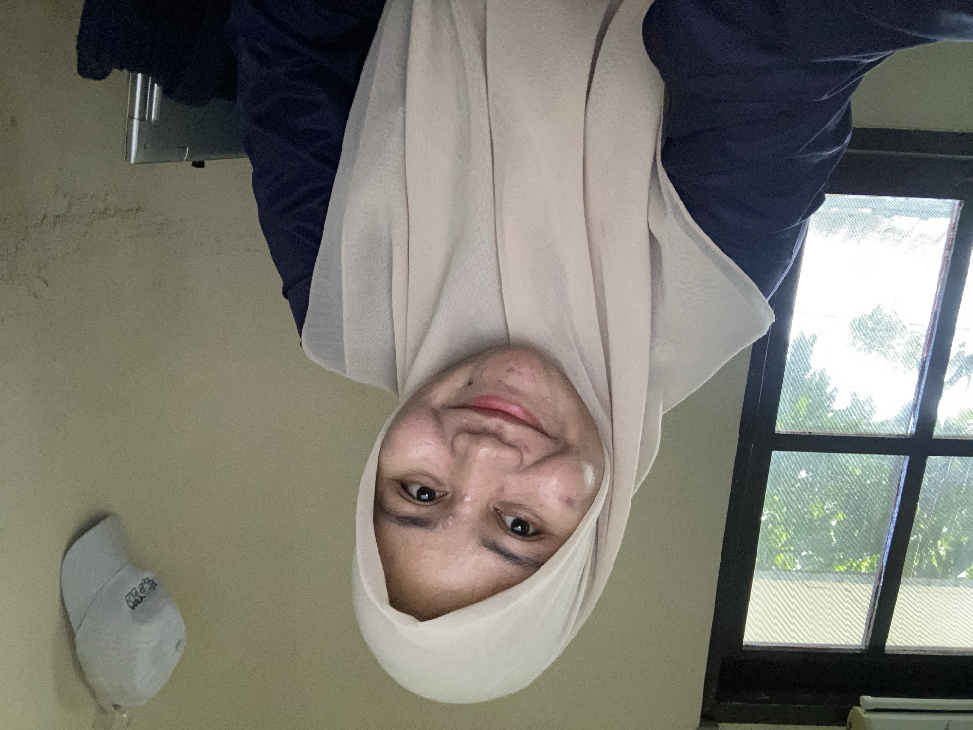 A medium shot shows a young woman wearing a cream-colored hijab and a dark blue top. She is looking directly at the camera and smiling. Her skin has some blemishes, and there is a small mole on her chin. Her head is tilted slightly to her left. The background consists of a wall and a window with light filtering through. A white baseball cap with a logo is hanging on the wall to the right of the woman. An air conditioner unit is visible above the window. The scene appears to be indoors, possibly in a residential setting in Banjarbaru, Indonesia.