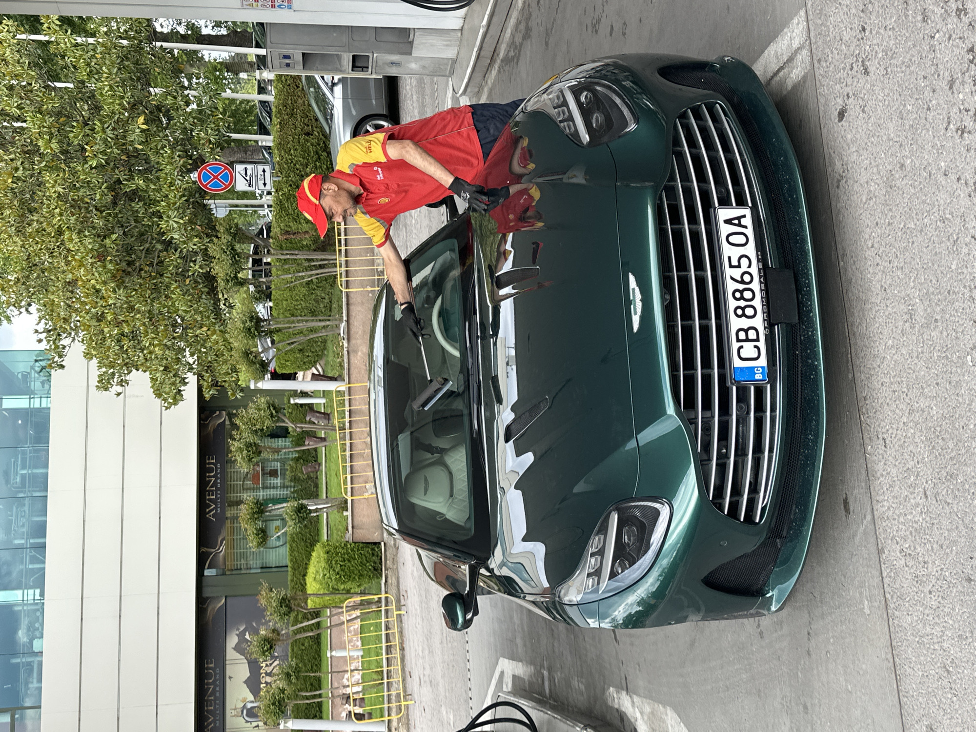 A man in a red and yellow uniform, wearing a red cap and black gloves, is washing the windshield of a green Aston Martin. The car has a Bulgarian license plate that reads "CB 8865 OA". The location appears to be a gas station in front of a building with the inscription "AVENUE MULTI BRAND" on the facade, situated near a tree and a street sign that indicates "no parking". The photograph was taken in Varna, Bulgaria.