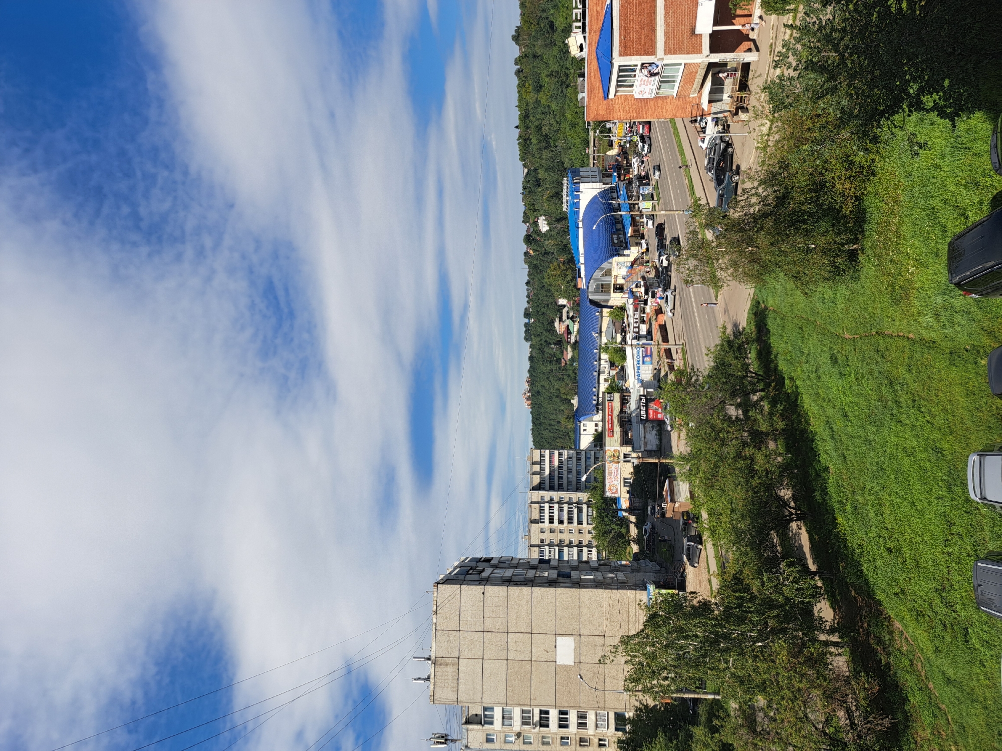 The image depicts a street scene in Irkutsk, Russia, on a clear, sunny day with scattered clouds. In the foreground, a steep, grassy embankment is visible, with several parked cars at its base. A two-lane road runs horizontally across the middle ground, with a moderate amount of vehicle traffic, including cars, vans, and a bus.
Several commercial buildings line the road. To the left, a large, grey, multi-story apartment building dominates the view, with several other similar buildings behind it. A complex with a blue, curved roof, likely a shopping center or market, is prominent in the center of the frame, with various businesses and advertisements visible. To the right, a brick building with a blue roof and a white facade on the upper level appears to be another commercial establishment.
The background features a dense, tree-covered hillside that rises behind the buildings. The overall impression is of a typical urban environment in a Russian city, with a mix of residential and commercial structures set against a natural backdrop. The presence of utility wires crisscrossing the sky suggests a developed infrastructure.