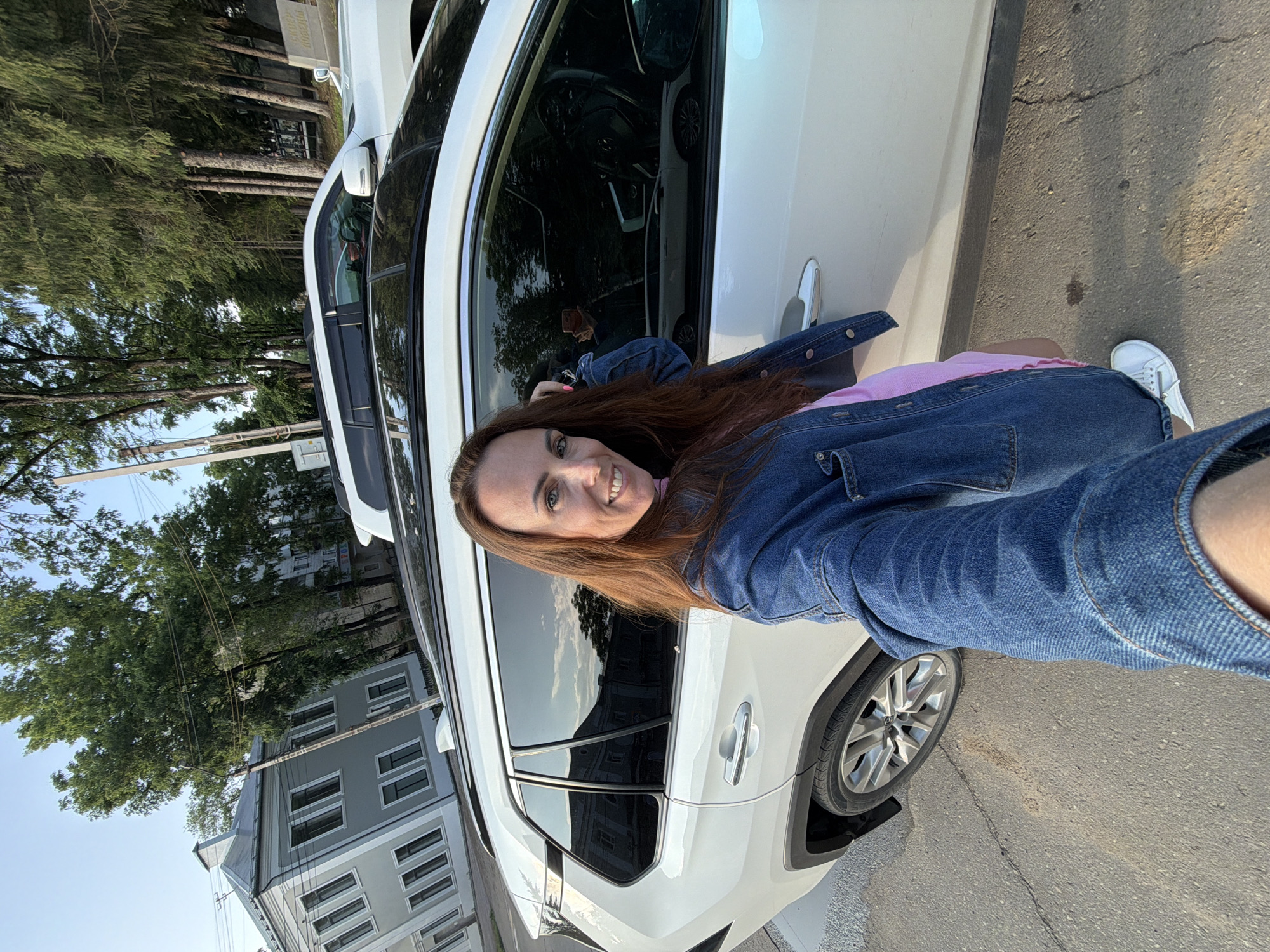 A woman with long brown hair, wearing a denim jacket and a pink dress, stands in front of a white SUV on a street in Birobidzhan, Russia. The woman is smiling and holding her phone up to take a selfie. The SUV's windows are tinted, and a reflection of another white car can be seen in the driver's side window. In the background, there are buildings and trees. The street is paved, and there is a sidewalk. The sky is clear and blue.
