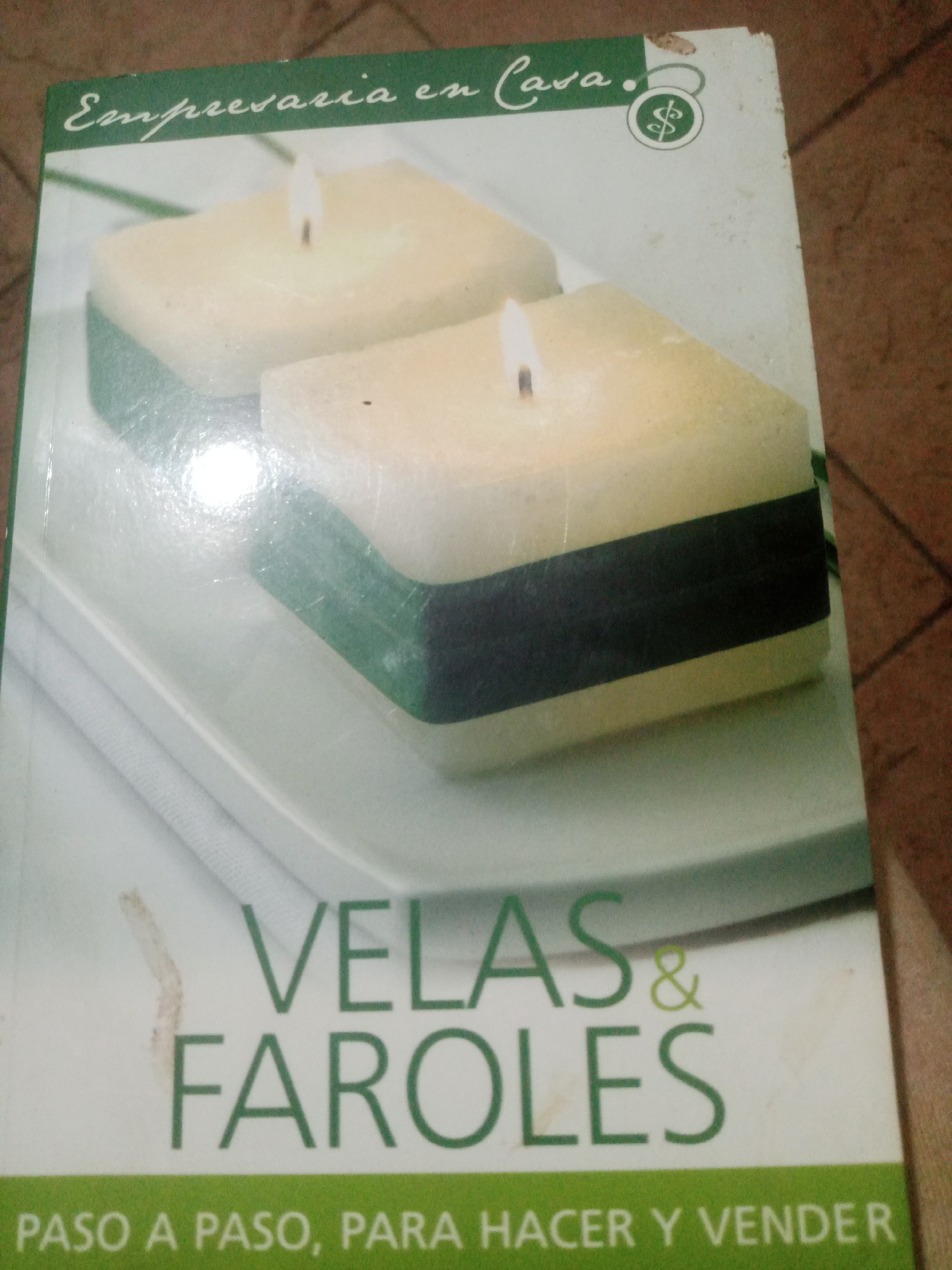 The image is a photograph of the cover of a book titled "Empresaria en Casa. Velas & Faroles. Paso a Paso, Para Hacer y Vender." The book cover features a close-up image of two decorative candles with lit wicks, resting on a white plate or tray. The candles appear to be made of a creamy-colored wax with a dark green band around their middle. The text on the cover is in Spanish. There is a dollar sign symbol inside a circle on the upper right side of the cover. The background is a blurry wooden surface. The overall lighting suggests the photograph was taken indoors. The location in the city of Santa Ana, Venezuela, is not discernible from the image itself. There are no people or interactions depicted in the image.