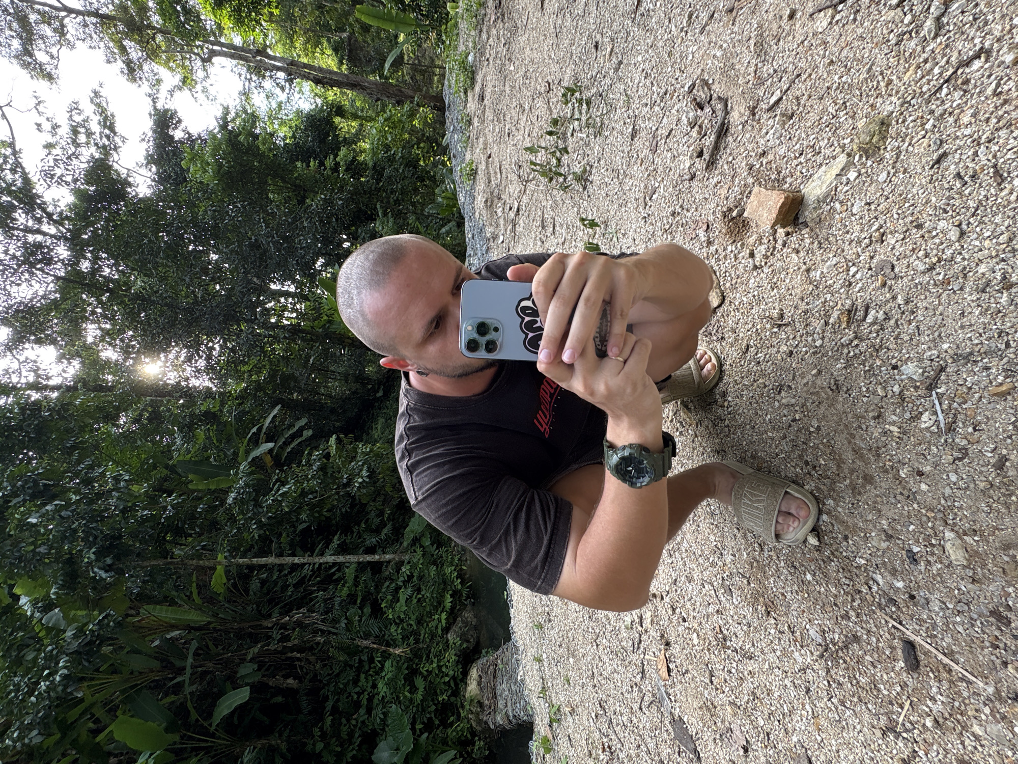 A bald man wearing a dark t-shirt and light-colored slides is squatting on a gravelly path in a lush, green forest. He is holding a light blue smartphone in both hands, appearing to take a picture or record a video. The man is wearing a dark green watch on his left wrist. Sunlight filters through the dense foliage of the trees overhead, creating dappled light on the ground. A small stream or body of water is visible to the left of the frame, bordered by rocks and vegetation. The environment suggests a natural, outdoor setting likely within or near a tropical forest, consistent with areas around Choeng Thale, Thailand.