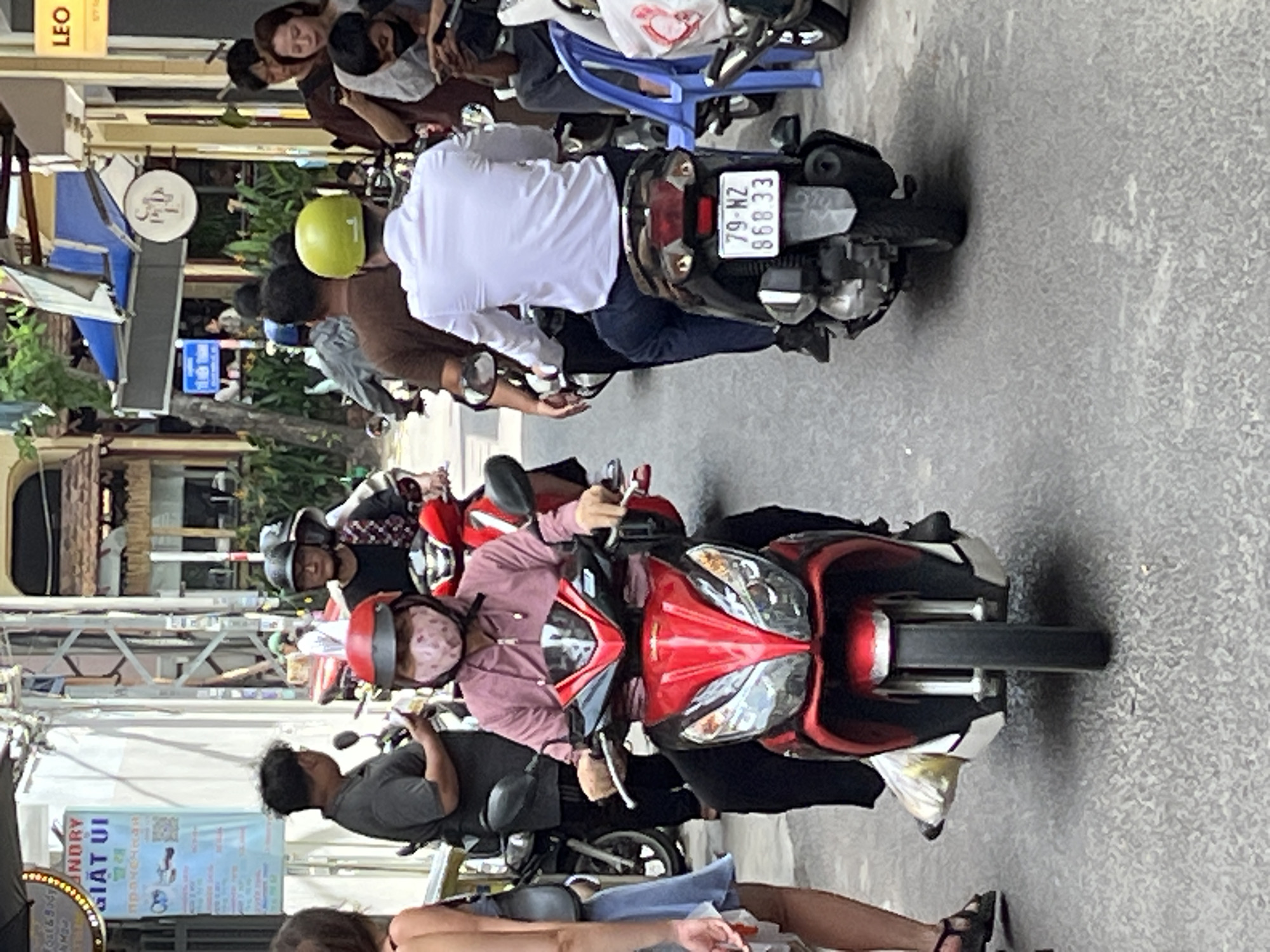 In Nha Trang Ward, Vietnam, on a street with buildings on both sides, a person wearing a pink jacket, black pants, and a patterned mask rides a red and black scooter. The scooter has a silver front and black handlebars, with the front wheel visible. The rider's hands are on the handlebars, and their feet are on the floorboard. Behind this scooter, another person wearing a white shirt and dark pants rides a black scooter with a yellow helmet. The license plate of this scooter reads "79-WZ 868.33". In the background, other people and scooters are visible, along with signs and shop fronts, suggesting a commercial or residential area. The ground is paved with asphalt.