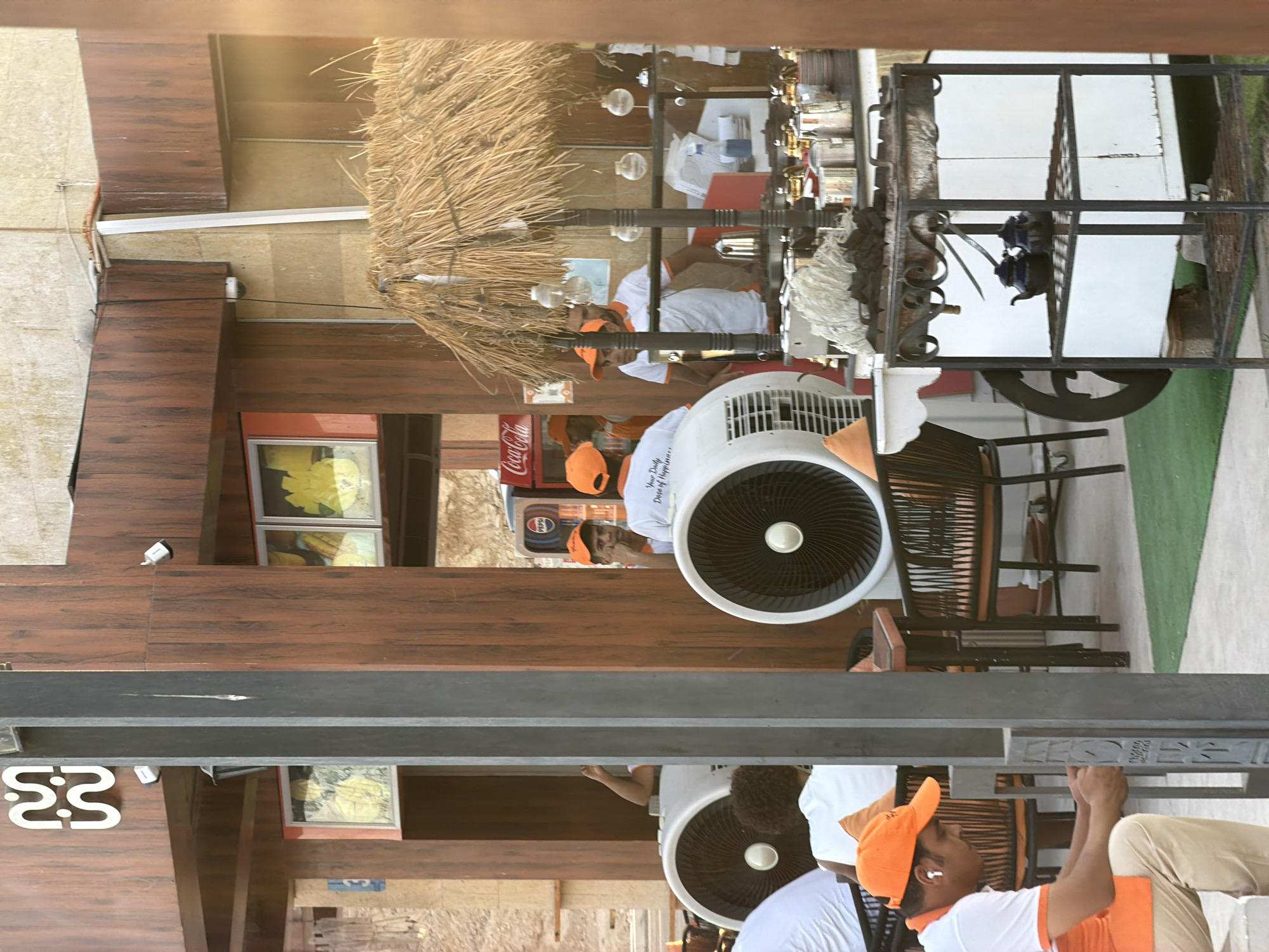 The image captures a scene outside a food establishment, likely a cafe or restaurant, in Sharm El Sheikh, Egypt. The architecture features wooden paneling and a rustic straw-thatched roof over a service cart. Several individuals, appearing to be staff members, are present. One person in the foreground, wearing an orange cap and white shirt, is seated and appears to be engaged in a task near a gray column. Other staff members in similar uniforms are visible behind a counter, some interacting with each other or attending to their duties. Large industrial fans are placed around the seating area, suggesting a warm climate. The service cart is laden with items, including teapots and what appears to be charcoal, indicating food or beverage preparation. A Coca-Cola dispenser is also visible in the background. The overall environment suggests an outdoor dining or service area, possibly a tourist-oriented location given the context of Sharm El Sheikh.
