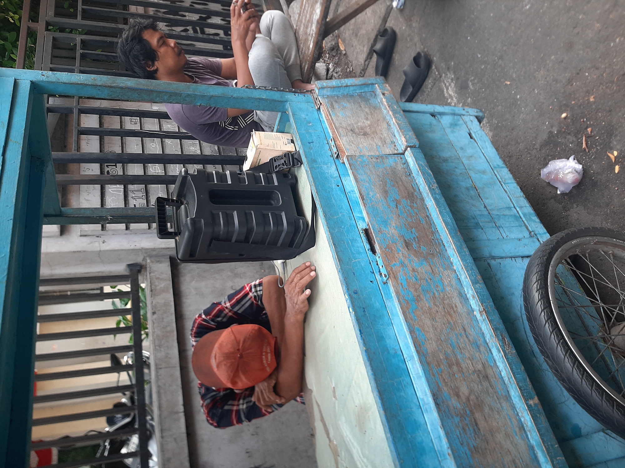 A man in a red and blue plaid shirt and an orange baseball cap sleeps with his head down on a light blue wooden counter of a mobile cart in Central Jakarta, Indonesia. The cart, also painted blue with weathered wood accents, has a black speaker on the counter near the sleeping man. Another man, wearing a dark purple t-shirt and grey pants, sits on a wooden bench to the right of the cart, looking down at his hands, possibly holding a phone. A small white box, labeled "PROTECTIVE MASK," and a black buckle are on the counter. The ground is asphalt with some debris, and a black bicycle wheel is visible on the left side of the cart. A metal fence with vertical bars is behind the cart.
