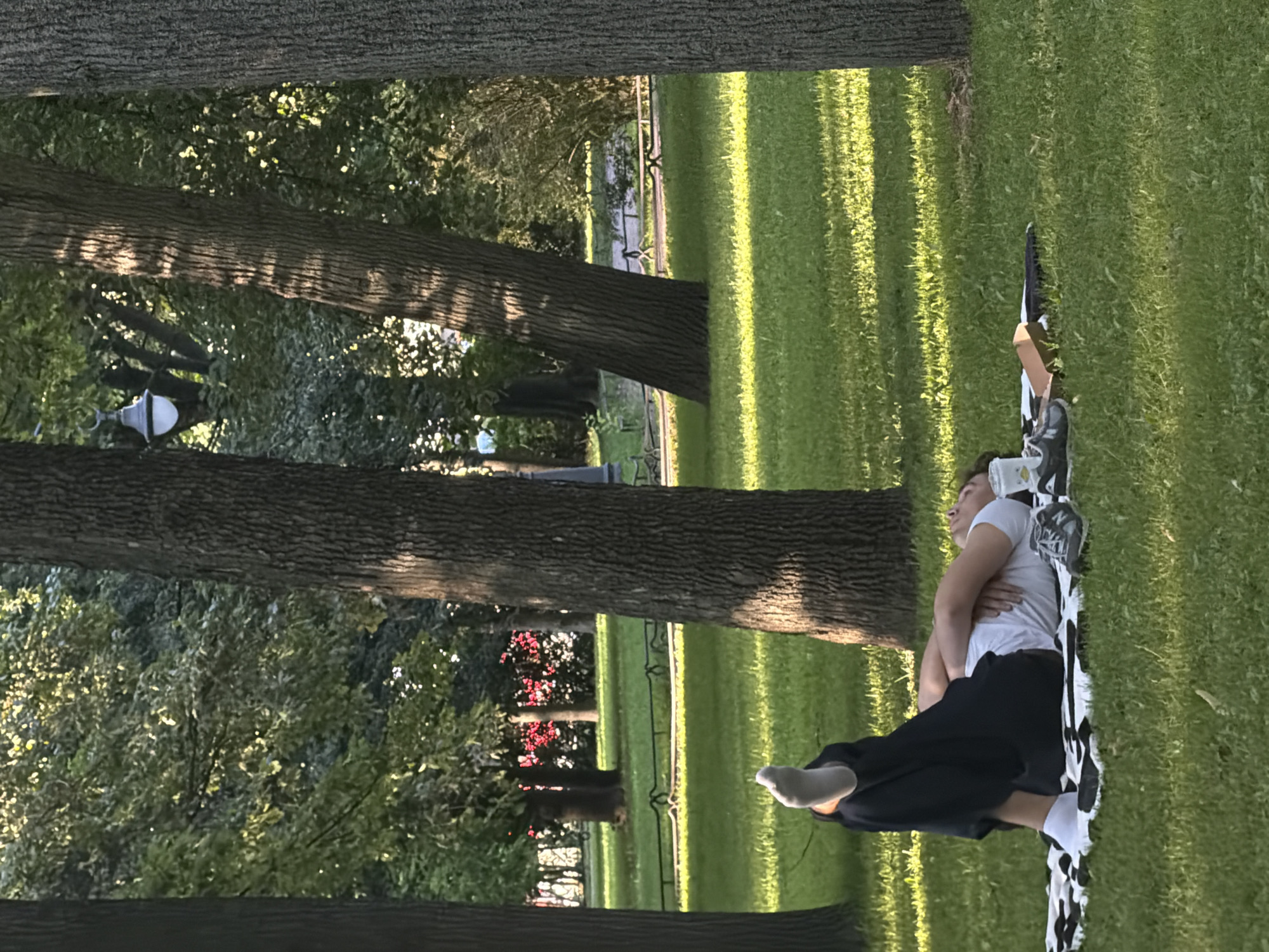 A young person is lying on their back on a black and white patterned blanket on a grassy lawn in a park in Saint Petersburg, Russia. The person is wearing a white t-shirt, dark pants, and white socks. Their legs are bent and raised, and their arms are crossed over their chest. Next to the person are a white cup, a cardboard box, and a pair of dark sneakers. The park features several large trees with thick trunks, casting shadows on the grass. In the background, there are more trees, some with green foliage, and a glimpse of red flowers. A lamppost is visible among the trees in the distance. The overall scene depicts a relaxed moment in a park on a sunny day.
