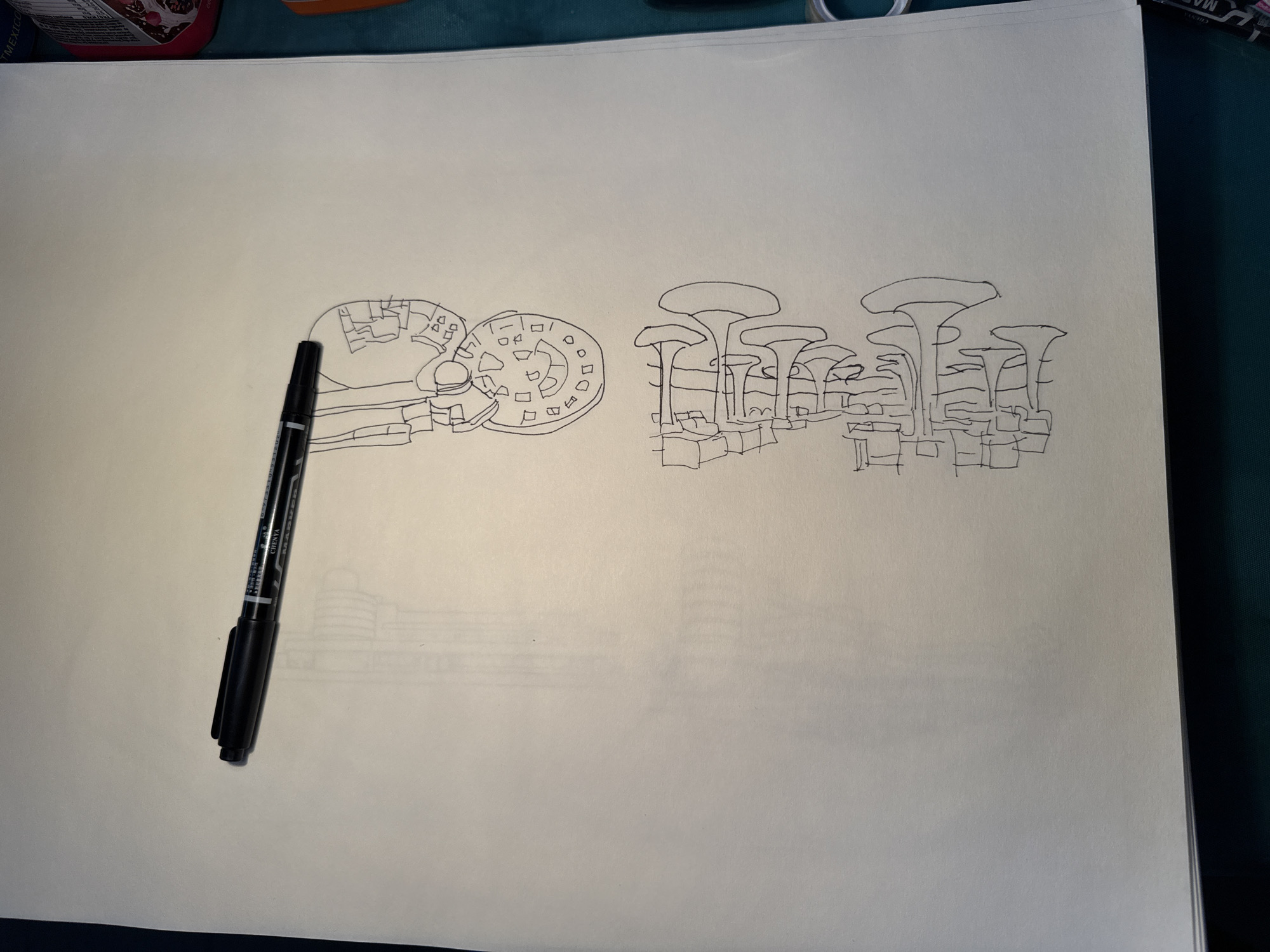 A hand-drawn sketch in black ink on a white paper. The sketch depicts two architectural elements: a stylized representation of the Fort of Guadalupe in Puebla, Mexico, with its circular ramparts and interior structures, and a group of mushroom-shaped structures, reminiscent of the Metropol Parasol in Seville, Spain, arranged atop a series of blocky bases. A black marker rests on the paper near the Fort of Guadalupe drawing. Faint lines indicating more buildings are drawn under the Fort of Guadalupe drawing.
