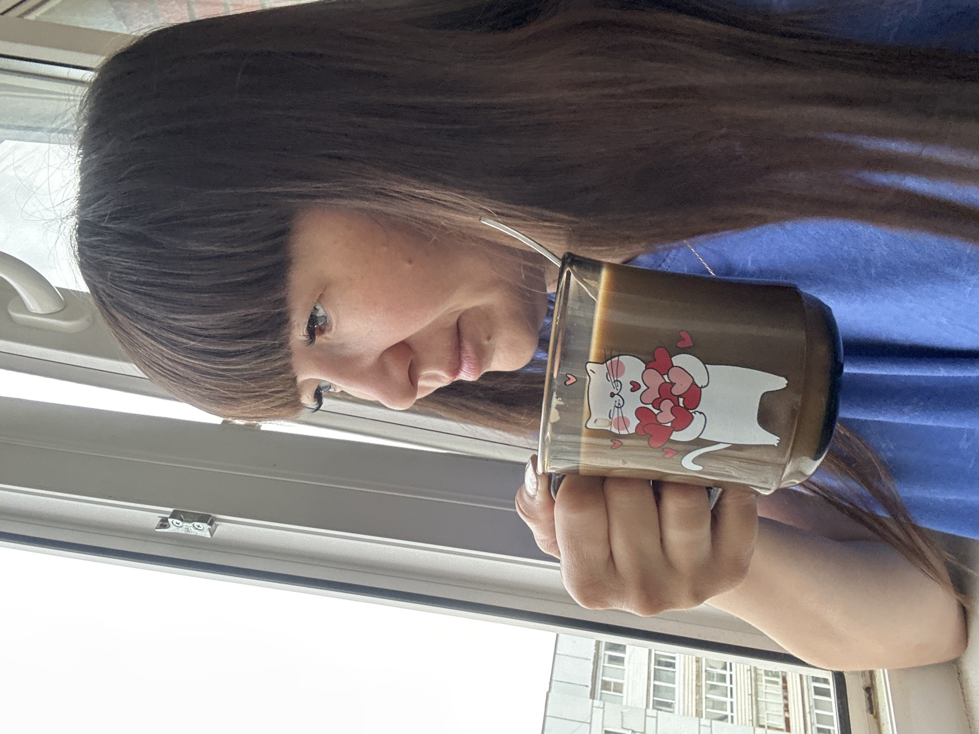The image portrays a young woman, likely in Chaikovsky, Russia, captured in a close-up, angled shot by a window. She has long, straight dark brown hair with bangs that obscure part of her forehead. Her face is partially visible, showing her right eye with dark eyelashes, her nose, and a slight hint of a smile on her mouth, indicating fair skin. She is wearing a blue garment, possibly a t-shirt or blouse.
She holds a clear glass mug in her right hand, her fingers adorned with light-colored nail polish. The mug prominently features a cartoon graphic of a white cat holding multiple red hearts. It contains a light brown beverage, likely coffee or a similar drink, with a metal straw inserted.
The woman is looking out of a window, which includes a white handle and metallic latches on its frame. Beyond the window, an urban or semi-urban environment is visible, featuring a multi-story building with numerous rectangular windows. The sky appears bright white and overcast, indicating daytime conditions without direct sunlight.
The scene conveys a relaxed, contemplative atmosphere, as the woman appears to be enjoying her drink while gazing outside. The most notable graphic detail is the white cat with red hearts on the mug.
