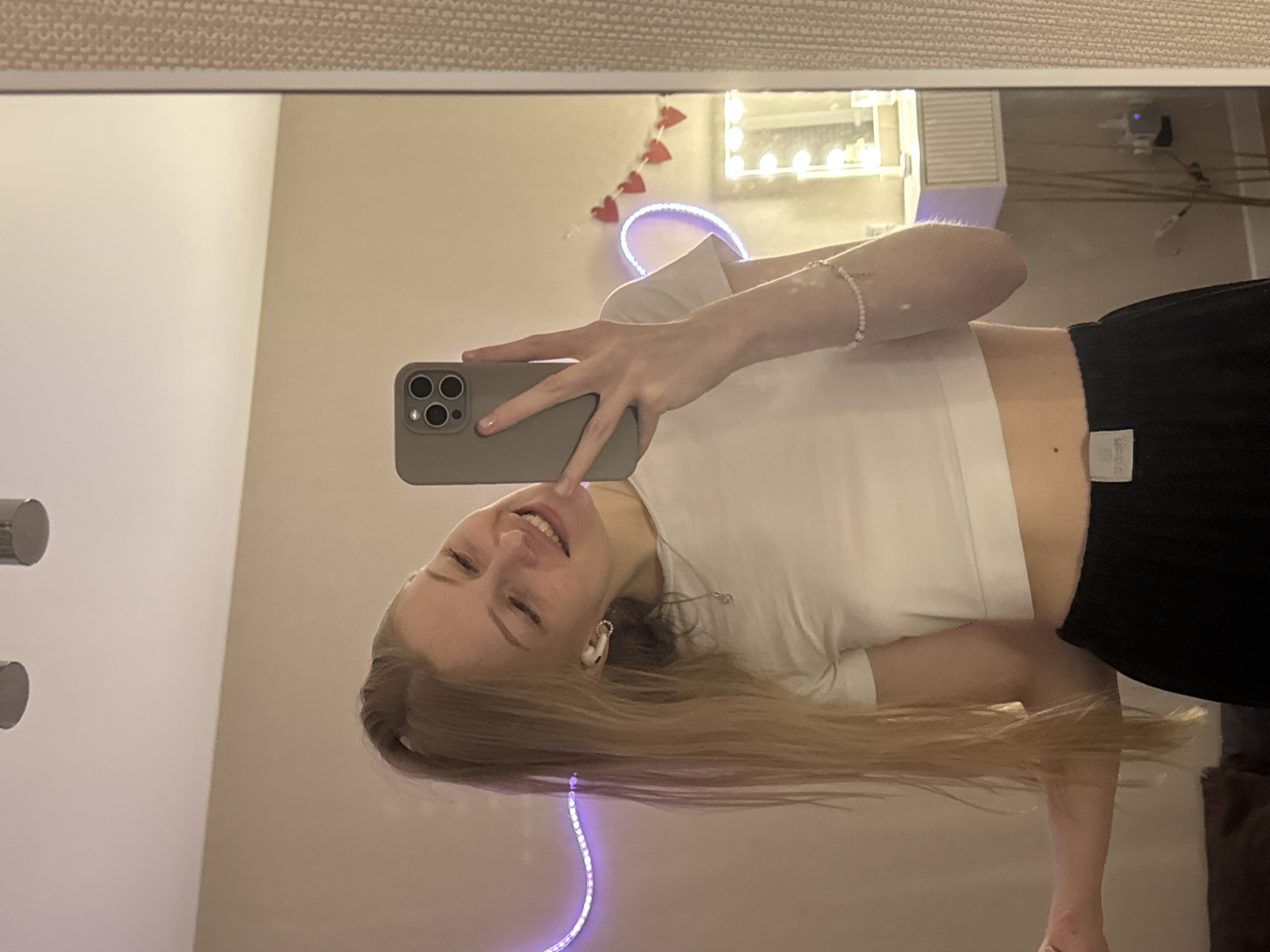 A young woman is captured in a mirror selfie, smiling brightly in an indoor setting in Perm, Russia. She holds a grey-cased smartphone with a triple camera lens setup, angled to take the photo. Her long, straight blonde hair flows past her shoulders. She is wearing a white, short-sleeved crop top and black bottoms, with a small white tag visible on the waistband of the latter. A light-colored beaded bracelet adorns her right wrist, and a white earbud is visible in her left ear.
The reflection reveals a cozy room ambiance. A string of red heart-shaped decorations hangs on the wall behind her. A lighted vanity mirror with multiple bulbs and a glowing purple LED strip add to the room's decor. The large mirror is framed by a light-colored, textured border at the top. The overall mood is cheerful, emphasized by the woman's wide smile and relaxed posture. The lighting suggests an indoor setting, likely with artificial lights contributing to the bright scene.