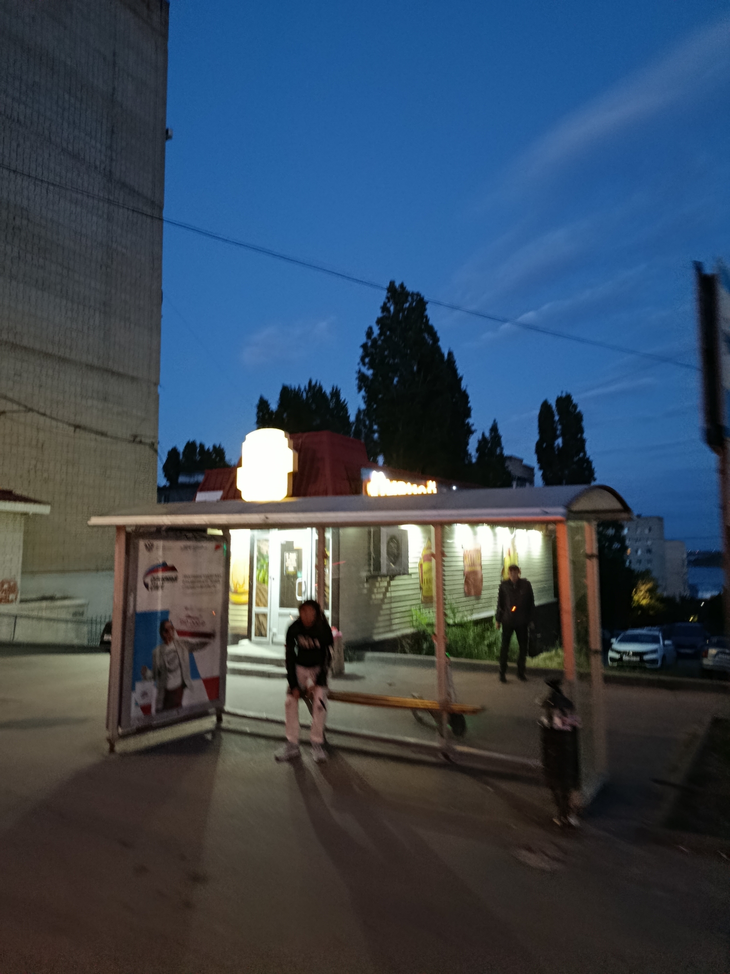 The image shows a bus stop in Saratov, Russia, at dusk. A kiosk with a bright light on top is located behind the bus stop shelter. A person is sitting on a bench inside the shelter, facing away from the camera, wearing a dark hoodie and light-colored pants. Another person is standing in front of the kiosk, wearing a dark jacket and pants. Cars are parked on the street to the right of the bus stop. The sky is dark blue with some faint clouds. A tall building is visible to the left of the scene, and trees are in the background.