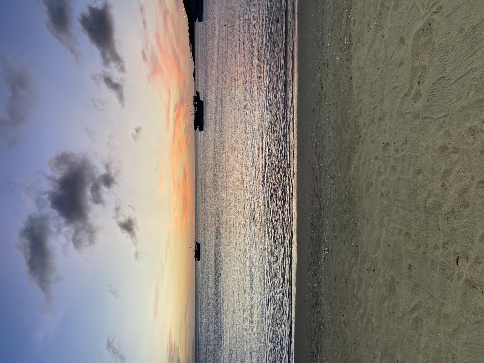 The image displays a tranquil coastal scene captured during either sunrise or sunset in Juan Griego, Venezuela. The landscape is horizontally divided into three main sections: the sky, the sea, and a sandy beach.
The sky, occupying the left portion of the image, showcases a beautiful gradient of colors, transitioning from a soft blue on the far left to warm shades of yellow, orange, and pink towards the right, indicative of a transitional time of day. Several dark, scattered clouds are visible against this colorful backdrop.
The central part of the image features a calm body of water, likely the Caribbean Sea, which gently reflects the sky's soft light. The water's surface is textured with small, shimmering ripples. Two small, dark boats are visible in silhouette on the water; one is more prominent in the mid-ground, and another appears further in the distance. A faint, dark outline of land can be seen on the horizon beyond the boats.
To the right, a wide stretch of sandy beach fills the foreground. The sand displays subtle textures, including faint wave patterns and marks, possibly footprints. No people are visible, and there is no discernible text. The overall atmosphere is one of serene natural beauty and peaceful solitude at the coast of Juan Griego.