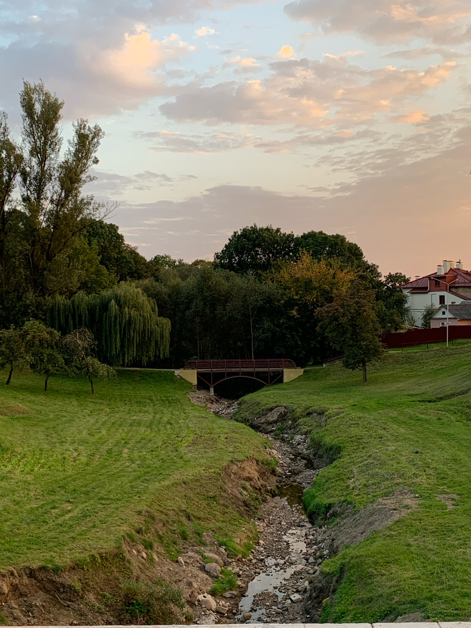 This is a vertical, eye-level, medium shot of a park in Hrodna, Belarus, on a cloudy evening. The sky is filled with wispy, pastel-colored clouds, illuminated by the setting sun. A gentle slope of lush green grass occupies the foreground and mid-ground, with a dry, rocky creek bed cutting through it. A small, arched wooden bridge spans the creek in the mid-ground. To the left of the bridge, a large weeping willow tree dominates the landscape, its branches cascading downwards. Behind the willow, a dense line of dark green trees forms a backdrop. Further to the right, beyond the trees, a section of a residential area with red-tiled roofs and white houses is visible, along with a dark red fence. No people or animals are present in the image.