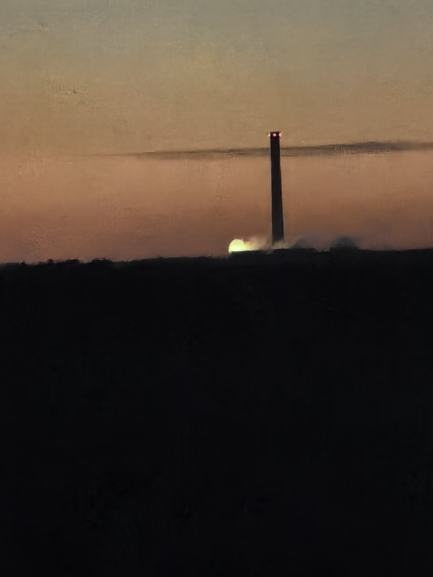 A tall, slender smokestack with red lights at its top stands against a hazy, orange-toned sky, likely during sunset or sunrise. A plume of light-colored gas or smoke emanates from the top of the smokestack. The lower half of the image is dominated by a dark, silhouetted landscape, likely a field or hilly terrain, obscuring any details on the ground. The overall impression is one of industrial activity occurring at dusk or dawn.