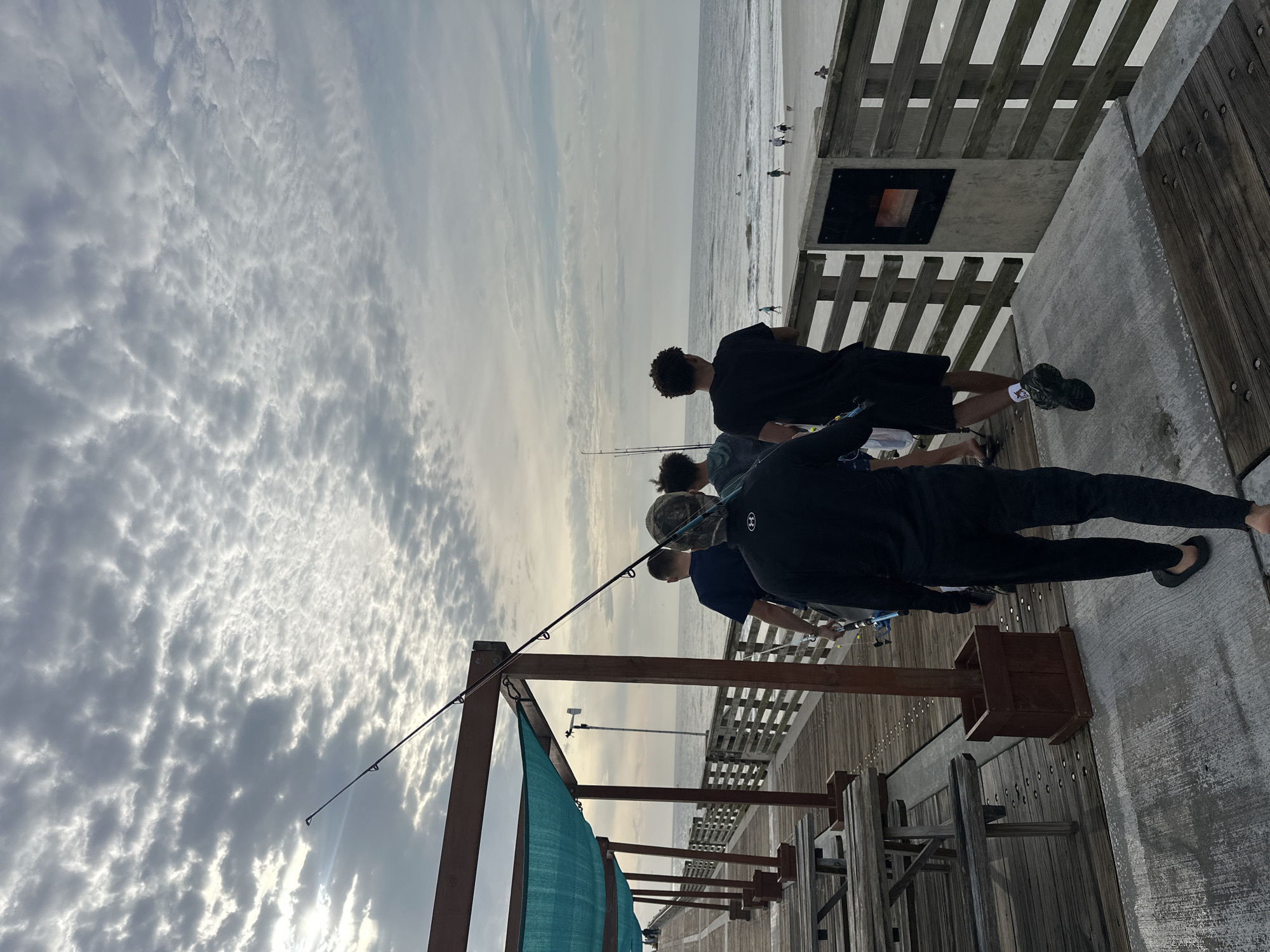 The image depicts a group of people fishing from a pier. The pier has wooden planks and railings. The sky is cloudy. In the distance, there's a beach with a few people on it. Fishing poles are visible, extending over the water. Based on the pier's architecture, location, and available landmarks visible in the photo, this scene is likely from Jacksonville Beach Fishing Pier located in Jacksonville Beach, Florida, United States of America.