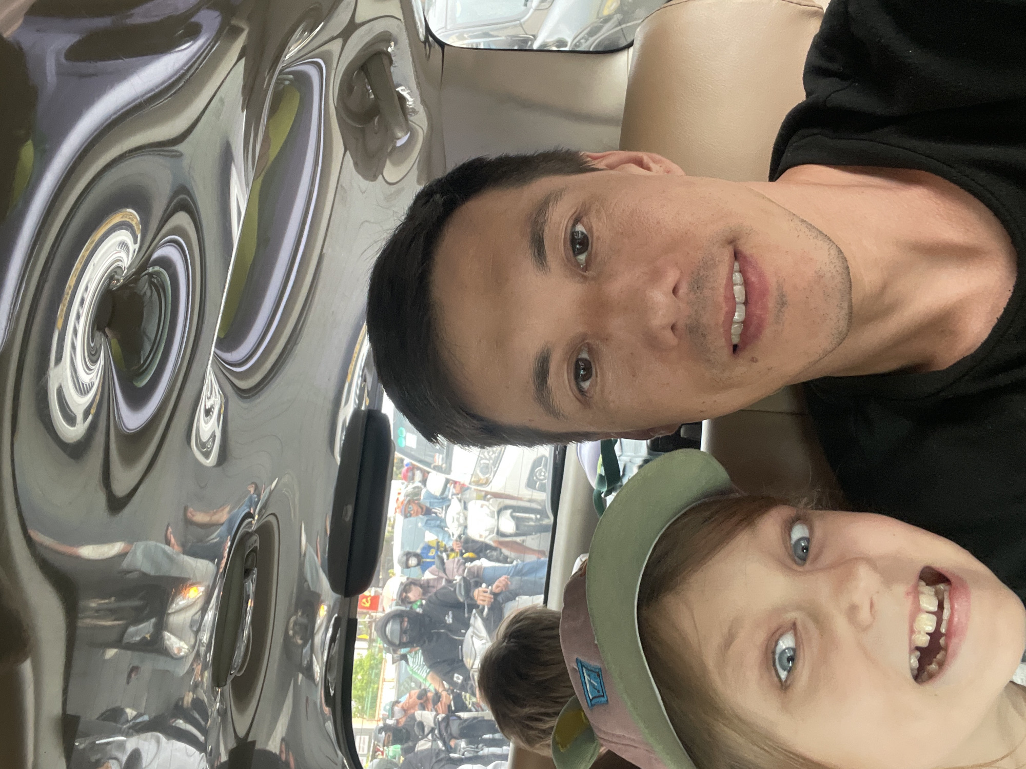 The image is a selfie taken from inside a vehicle in the city of Thủ Đức, Vietnam. A man and a child are visible in the foreground, with the man's face taking up the right side of the frame and the child's face on the left. The child is wearing a green and pink baseball cap. The background shows traffic, with many motorcycles and riders, suggesting a busy urban environment. The interior of the vehicle has a highly reflective ceiling with distorted reflections of the surroundings. The image appears to have been taken during the daytime.