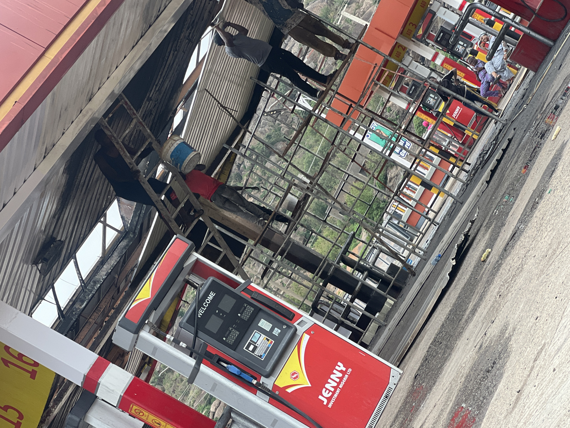 Here's a matter-of-fact description of the scene:
Location: Gura Top, Nigeria, a gas station featuring "JENNY INVESTMENT NIGERIA LTD" branding.
People: Several individuals are present. One is on a ladder holding a bucket. Two others are on a metal structure above the gas pumps; one appears to be working. More individuals are visible near the other gas pumps.
Objects: The scene includes gas pumps, a metal structure, a ladder, buckets, corrugated roofing, and road surface with apparent tire marks. The gas pumps have "WELCOME" displayed on them. Signage with numbers is visible.
Actions: Workers are actively engaged in maintenance or repair work on the gas station's structure. Customers are at the other pumps, possibly refueling.
Environment: The backdrop includes hilly or mountainous terrain. The scene shows evidence of possible construction or renovation underway at the gas station.