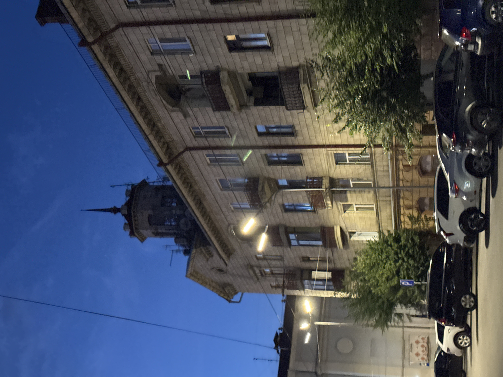The image depicts a twilight scene in Chernivtsi, Ukraine, focusing on a multi-story building with an ornate facade and a distinct turret. The building is illuminated by streetlights, casting a warm glow on its windows and balconies. Several cars are parked along the street in front of the building, including a black minivan, a white hatchback, and a dark SUV. Trees with dense foliage line the street, partially obscuring the lower levels of the building. The sky is a deep blue, indicating the transition from day to night. The overall atmosphere is calm and urban.