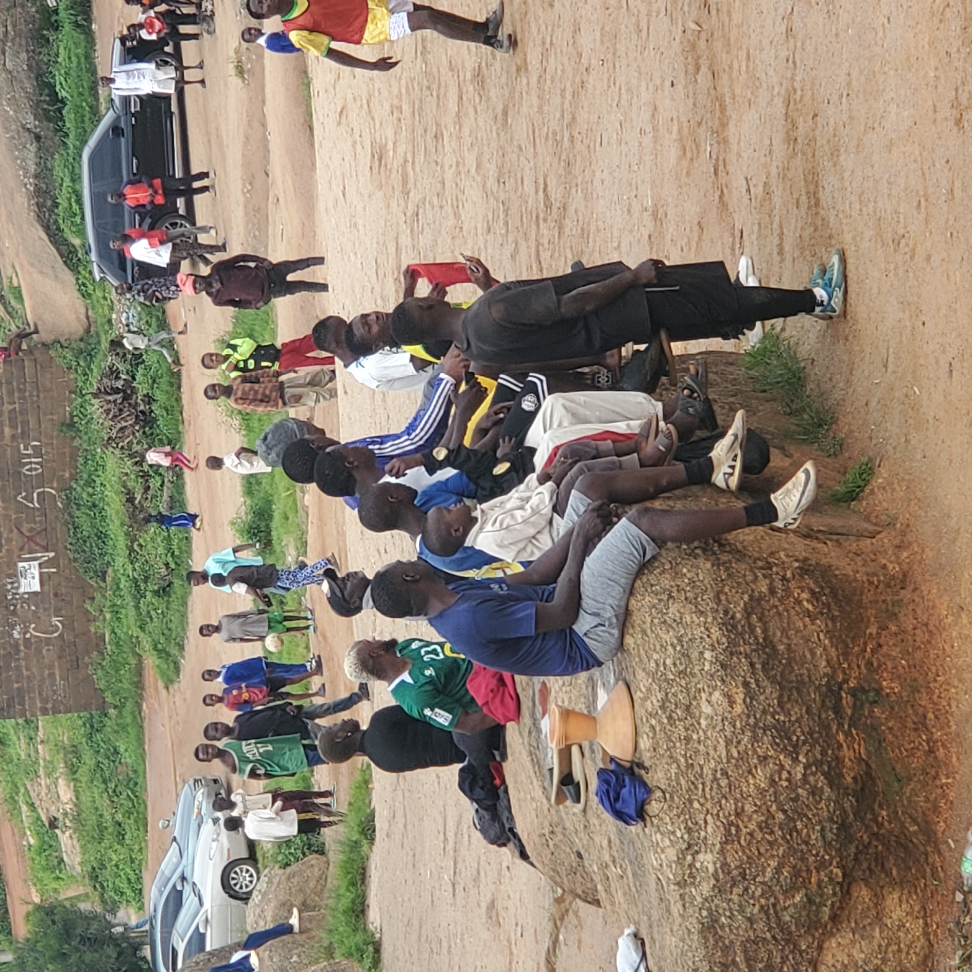 The image depicts an outdoor scene in a dusty, unpaved area with scattered rocks and vegetation. A group of people, predominantly young men and boys, are present. Some are sitting on a large rock formation, while others are standing or walking. There is a dark-colored SUV parked in the background, along with two cars. The people are casually dressed, and some appear to be engaged in a group activity, possibly related to sports given the presence of a soccer ball. The overall impression is of a community gathering in a somewhat informal, possibly rural or semi-urban setting.