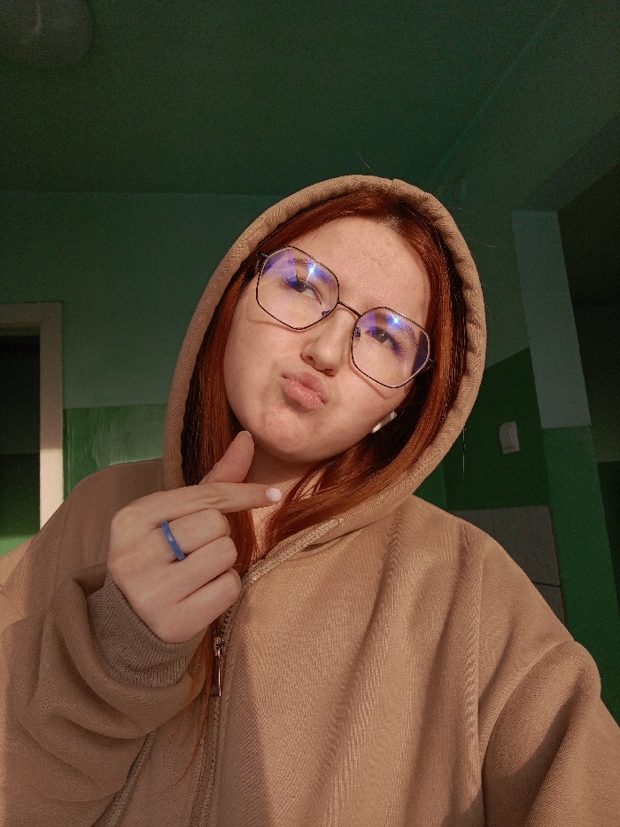 A young female subject is depicted in an indoor setting, identified as being in the city of Chaikovsky, Russia. She has reddish-brown hair and is wearing a light brown hooded sweatshirt with a subtle chevron pattern, partially zipped. She wears large, octagonal-framed eyeglasses with discernible reflections on the lenses, and a white earbud is visible in her right ear. Her lips are pursed, and her gaze is directed slightly upwards and to her left. Her right hand is raised, with the index finger extended and pointed towards the left side of her face. A blue, possibly silicone or plastic, ring is present on her right ring finger. The background features a wall painted in two horizontal sections: a muted teal upper portion and a darker green lower portion. A white door frame is visible on the left side of the frame, and a white rectangular object, consistent with a light switch or wall outlet, is affixed to the wall on the right. A faint circular light fixture is visible on the ceiling in the upper left corner.