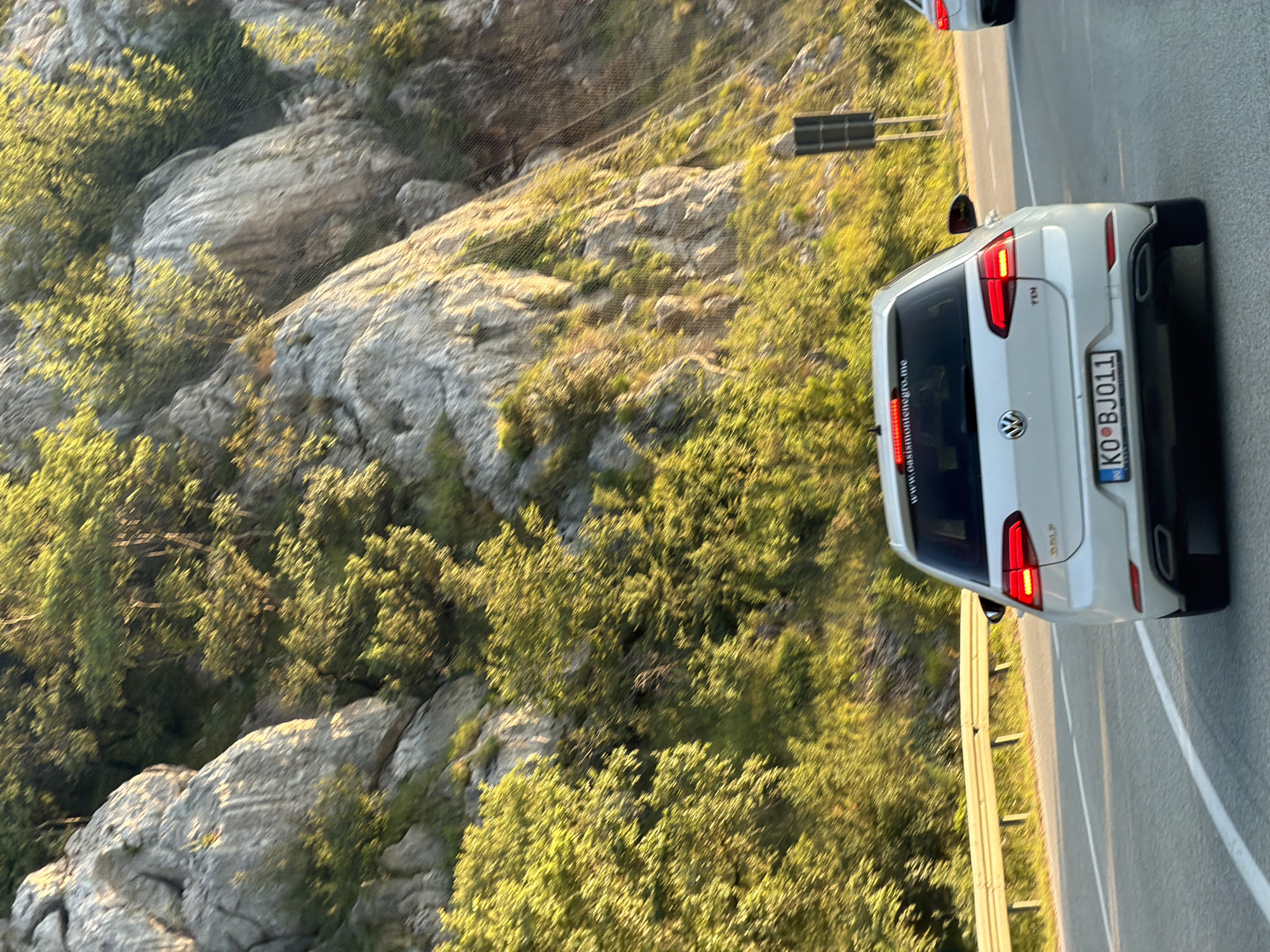 The image shows a white Volkswagen Golf TDi driving on a paved road. The car has a license plate that reads "KO BJ011" and a sticker on the rear window with the text "www.oasispmontenegro.me". The car is in motion, suggested by the blur of the surroundings and the car itself.
The road appears to be a scenic route, possibly a highway, flanked by a steep, rocky hillside covered in dense green vegetation. The sun is shining brightly, casting long shadows and illuminating the foliage with a warm, golden light. The sky is not visible in the frame. A metal guardrail runs along the side of the road.
In the background, another white car is partially visible, also in motion. A dark, rectangular traffic sign or utility box is positioned on the side of the road further up. The scene is likely taking place in a mountainous or hilly region. Based on the license plate and the website sticker, the location is suggested to be Markovići, Montenegro.
