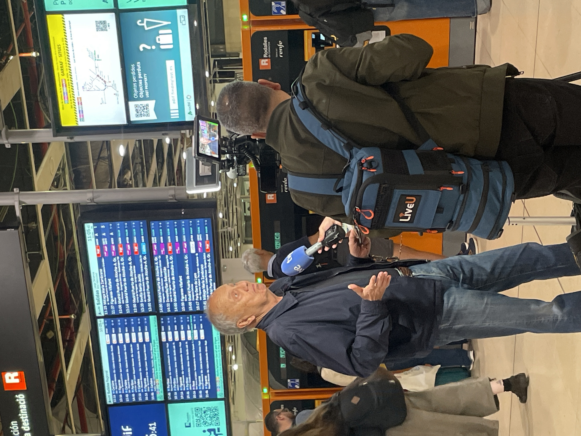 The image shows an interview taking place in what appears to be a train station, likely in Barcelona, Spain, given the signage.
A man is being interviewed, facing left, gesturing with his hands as he speaks into a microphone. A cameraman is recording the interview, his camera visible, with a small monitor attached. Another man stands behind the cameraman. A blue backpack with "LiveU" on it is visible.
In the background are departure boards displaying train schedules and platform information. There are also signs with information about lost property and other train-related information, including the Renfe logo. The lighting appears to be bright, common in public transport areas. The station seems relatively busy, although the focus is on the interview. A woman is seated to the left of the main subject.