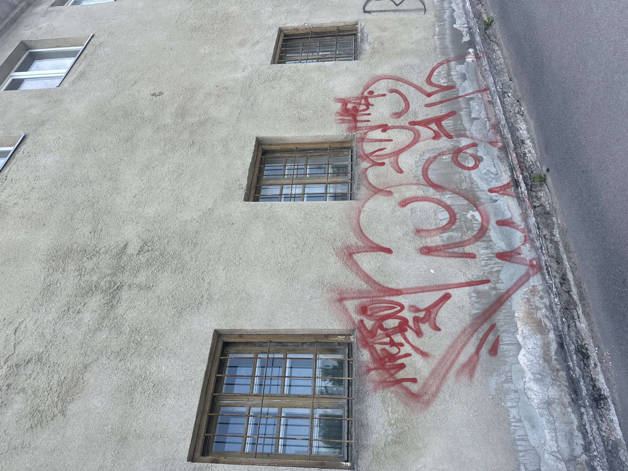 The image shows a section of a building's exterior wall in Tallinn, Estonia, featuring red graffiti spray-painted across the lower portion. The wall is constructed of a rough, light-colored stucco material. There are three windows visible on the wall, each with wooden frames and metal security bars. The windows reflect the sky and surrounding trees. A grey asphalt road runs along the bottom of the image, bordering the wall. The graffiti appears to be abstract lettering or symbols, with some portions smudged or faded. The building's architecture suggests it is likely an older structure.