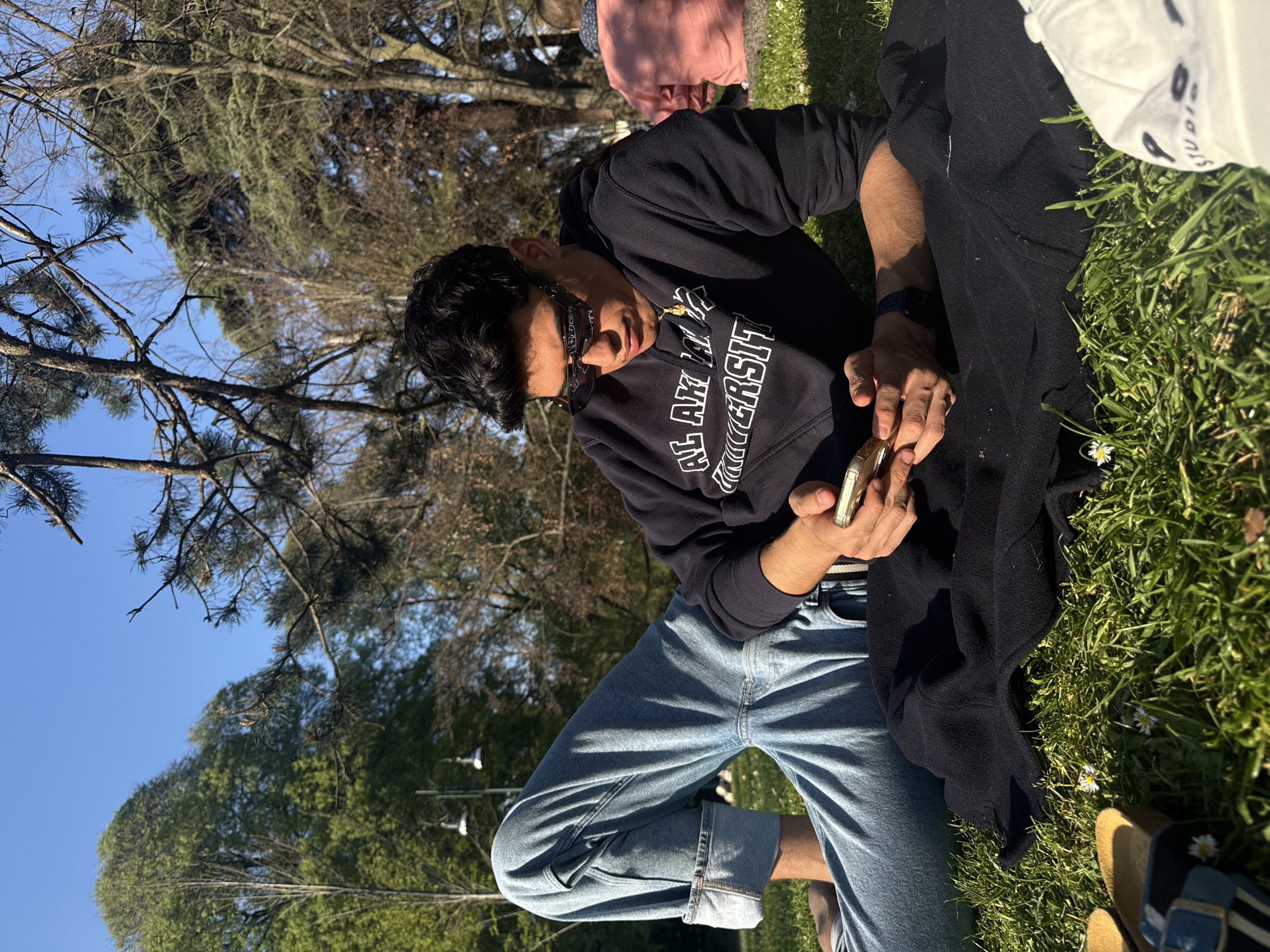 A young man is sitting on the grass in what appears to be a park. He is wearing a black sweatshirt with white lettering, light blue jeans, and sunglasses. He is holding a cell phone in his hands, appearing to examine it. A dark-colored jacket or blanket is partially covering his lap. The setting includes a grassy area, trees, and a clear blue sky. Other people are present in the background. Given the mention of Milan, Italy, without further information, I can only infer the location is a park in Milan.