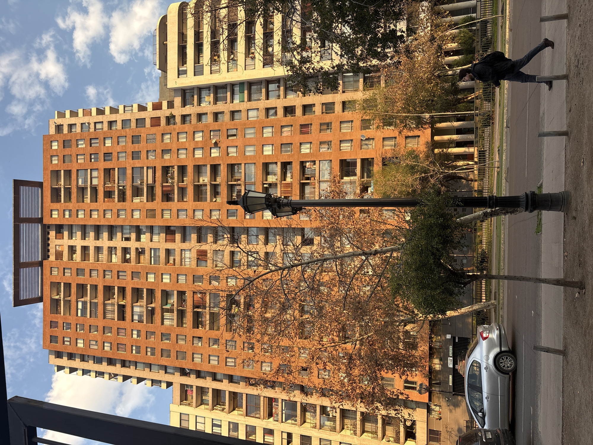This is a medium-angle, exterior, eye-level shot of a street scene in Santiago, Chile, featuring a tall, orange brick apartment building. The building has numerous windows and balconies, and is flanked by a lighter-colored building to its left and a white building with curved elements to its right. In the foreground, a silver sedan is parked on the left side of the street. A person wearing a dark jacket and jeans walks from right to left across a sidewalk, carrying a backpack. A black lamppost stands on the sidewalk. The street is lined with trees, some of which are bare or have sparse autumn foliage, casting long shadows in the late afternoon sun. The sky is partly cloudy. The image appears to be taken on a street like Avenida Providencia or a similar urban artery in Santiago.
