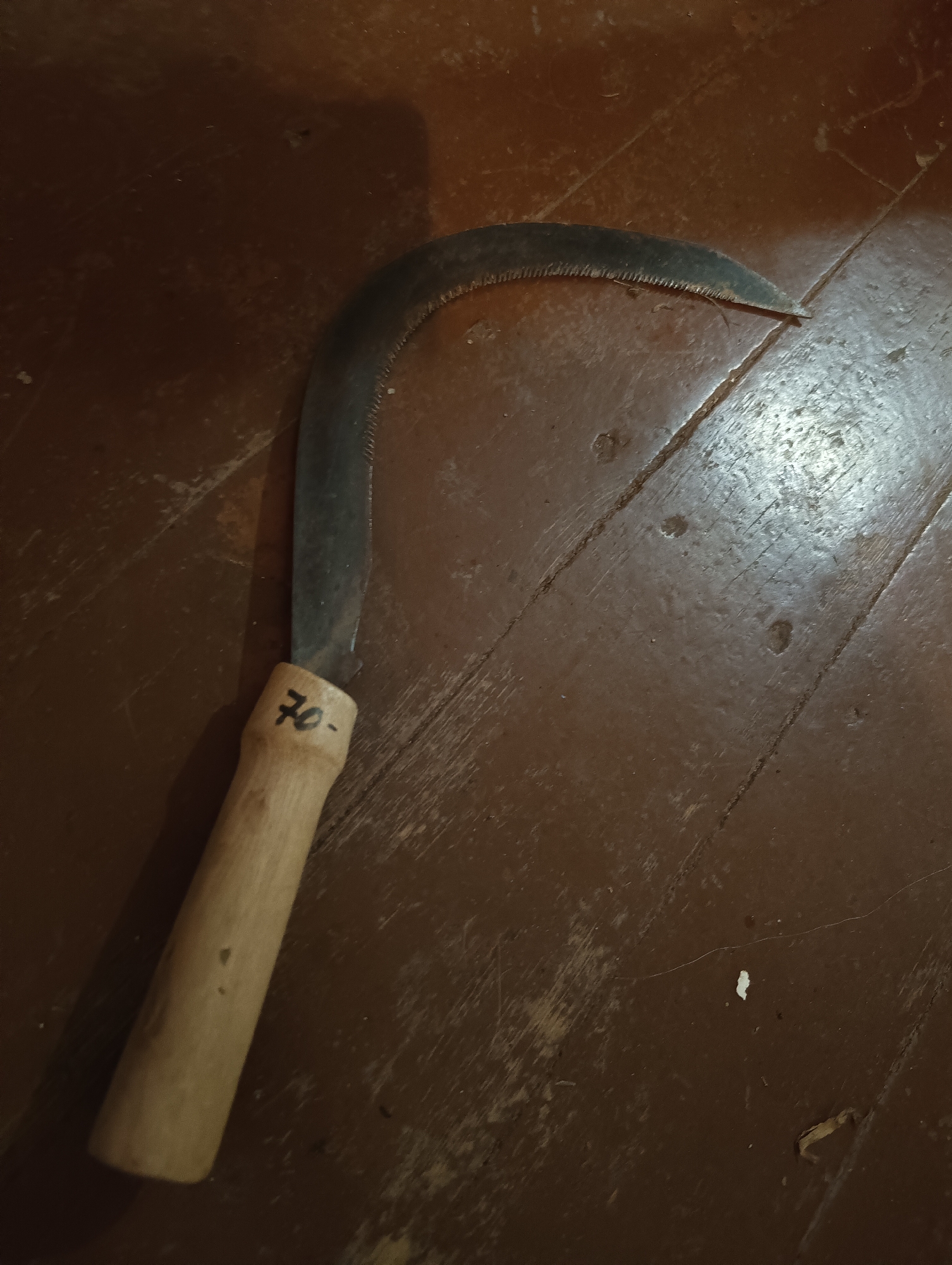 A metal sickle with a curved, serrated blade and a light-colored wooden handle is positioned diagonally on a dark reddish-brown painted wooden floor in Biliaivka, Odesa Oblast, Ukraine. The handle is marked with "70-" in black script. The floor exhibits visible plank seams, wear, scratches, and minor dust or debris. Natural or artificial light reflects off portions of the floor and the metal blade. No people or other significant objects are present.