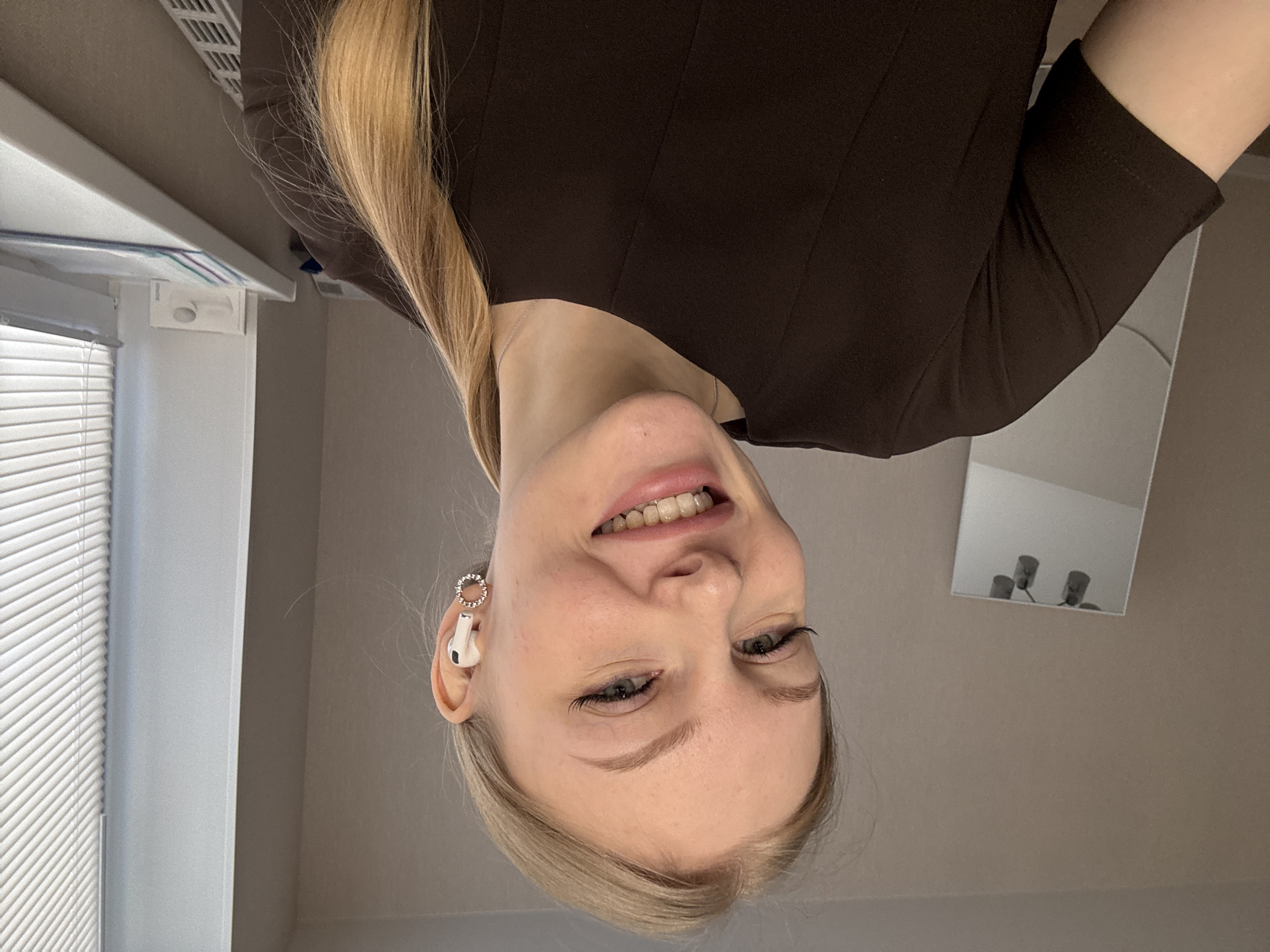 The image is an upside-down selfie of a fair-skinned woman with blonde hair, wearing a brown dress and an earring. She is smiling, and an AirPod is visible in her ear. In the background, there's a window with blinds and a mirror reflecting multiple items. Based on the individual's profile, it is plausible the image was taken in Perm, Russia.