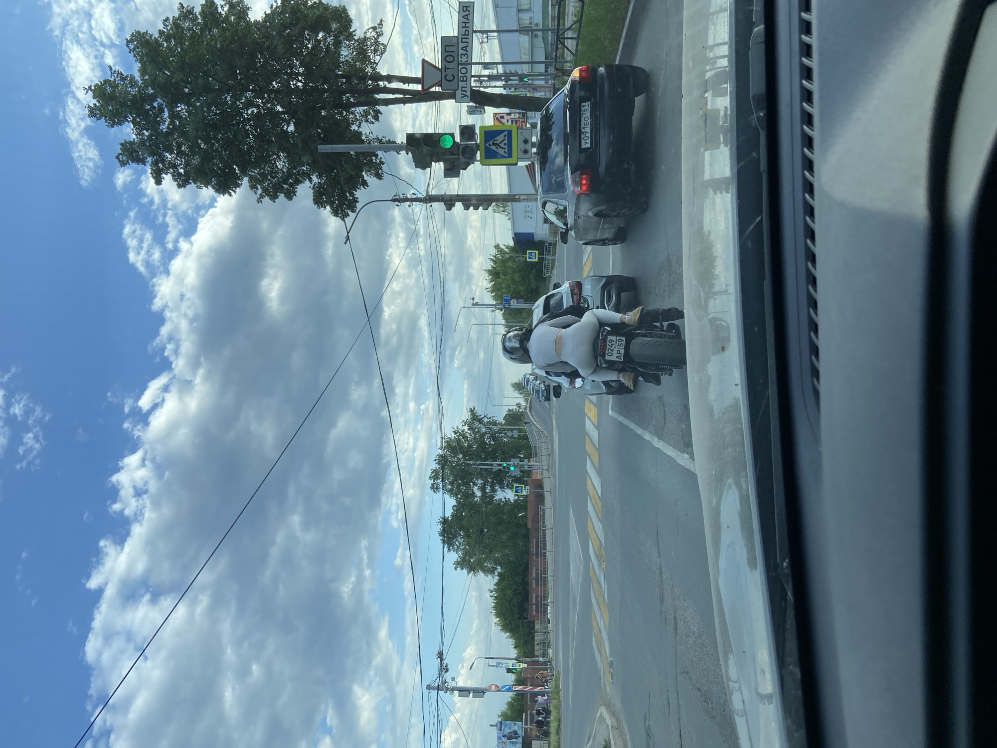 A view from inside a vehicle shows a street scene in Chaikovsky, Russia. The sky is partly cloudy. A black car with a license plate reading "Y091EO751" is stopped at a green traffic light on a road crossing. A motorcyclist wearing white clothing is positioned behind the car. A street sign with the word "CTON" is visible, along with "УЛ. ВОКЗАЛЬНАЯ". The street has a pedestrian crossing with yellow and white stripes. Trees and buildings line the road.
