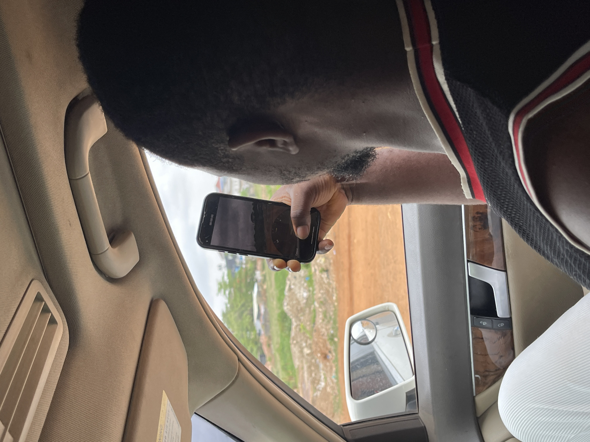 The image captures a first-person perspective from inside a vehicle, looking outwards. A person with dark skin and short, dark hair is partially visible on the left, holding a black smartphone in their right hand, seemingly looking out the window. Faint stubble or a short beard is visible on their cheek. They are wearing a dark athletic top with red and white stripes along the collar and shoulder. The phone screen is dark but shows a faint "00:00:00" which could indicate a camera app in standby or recording mode.
The vehicle's interior features a beige/tan upholstered ceiling and A-pillar, with a matching grab handle and an air vent. A sun visor with a "WARNING" label is also visible. The door panel has wood-grain trim with silver accents and two circular buttons, likely for window or mirror controls. Part of a black, textured steering wheel is seen on the far right, next to a white garment, possibly a passenger's leg or clothing.
Outside the vehicle, a reddish-brown dirt or unpaved road is prominent, flanked by green vegetation and some distant structures, suggesting an outdoor or semi-urban setting. A white side-view mirror with a small convex mirror attachment reflects a blurry scene. The sky appears bright and overcast, indicating daytime. The scene is consistent with typical road environments in places like Abuja, Nigeria.