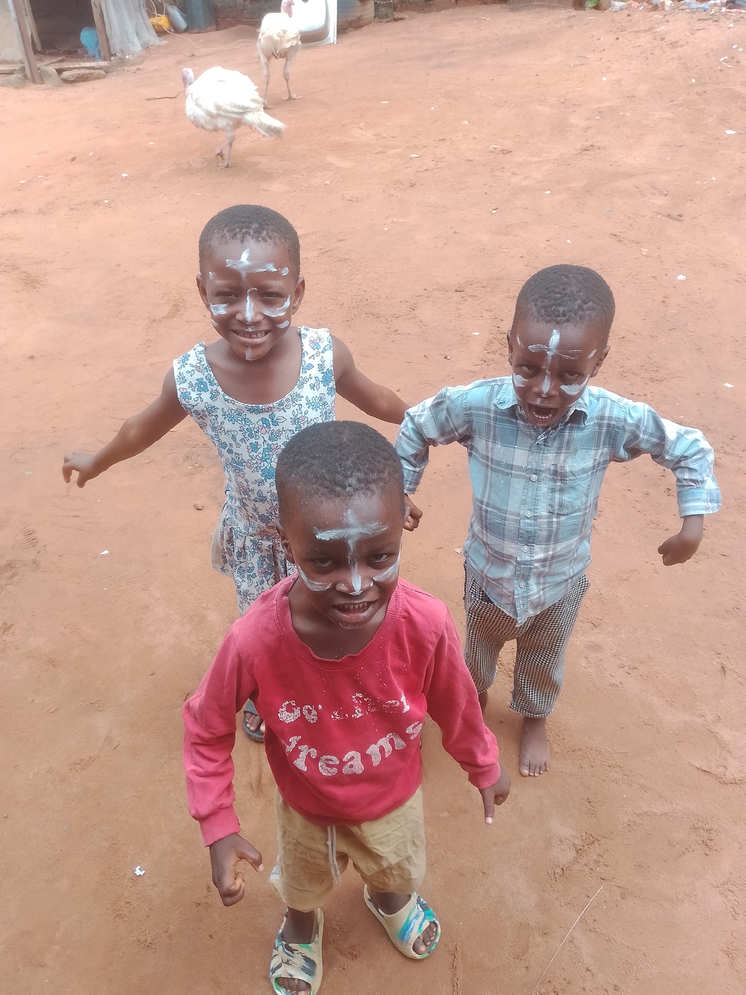 Three children with painted faces stand on a reddish-brown dirt ground in an outdoor setting. The child in the foreground wears a red long-sleeve shirt and beige shorts, with multicolored sandals. Behind him to the left is a child in a floral dress, and to the right is a child in a plaid shirt and checkered pants, barefoot. In the background, two white turkeys are visible. A structure with white netting is partially seen in the upper left corner of the frame. The location is Unknown, Unknown.