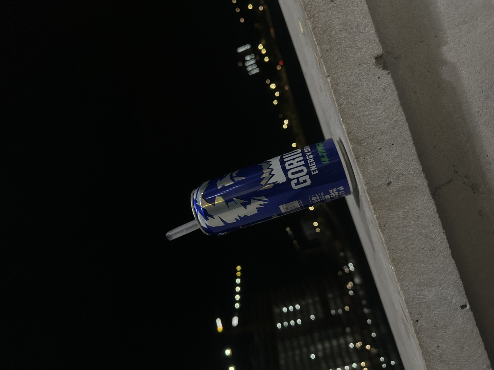 The image shows a blue Gorilla Energy Drink can with a silver and blue jagged pattern. A clear straw is inserted into the can. The can is sitting on a concrete ledge. In the background, there is a night cityscape with blurred lights indicating buildings. The location is likely a city, but specifics are not identifiable. The composition and lighting suggest a late-night or early-morning setting.