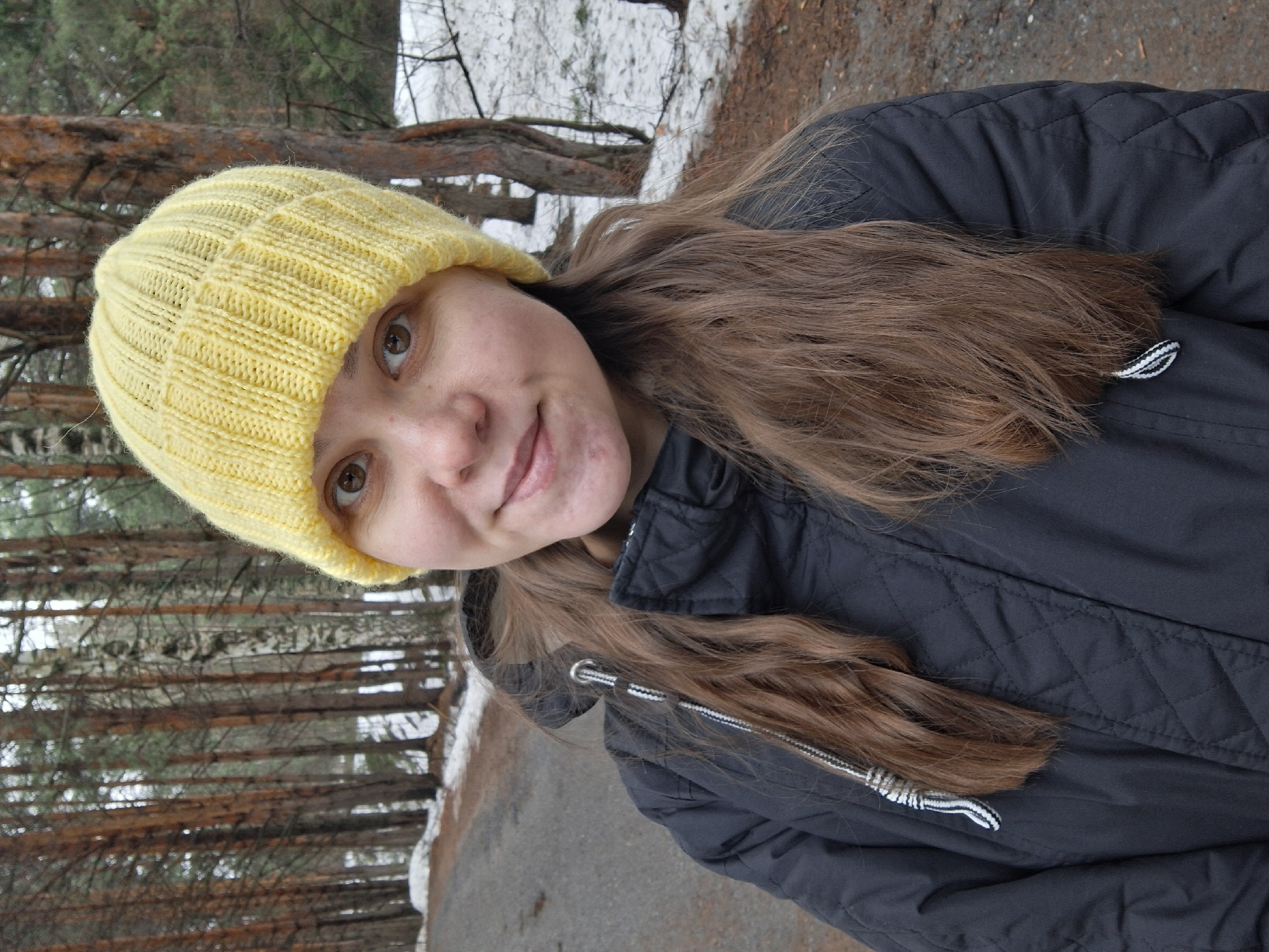 The image is a medium shot of a woman in what appears to be a park or wooded area. She is wearing a bright yellow knitted hat and a dark-colored, possibly quilted, jacket. She has light brown hair. In the background are trees, some covered in snow. The presence of snow suggests the image was taken during winter or early spring. The location is stated as Chaikovsky, Russia. The woman appears to be looking at the camera. There is a paved path or road visible in the lower part of the image.