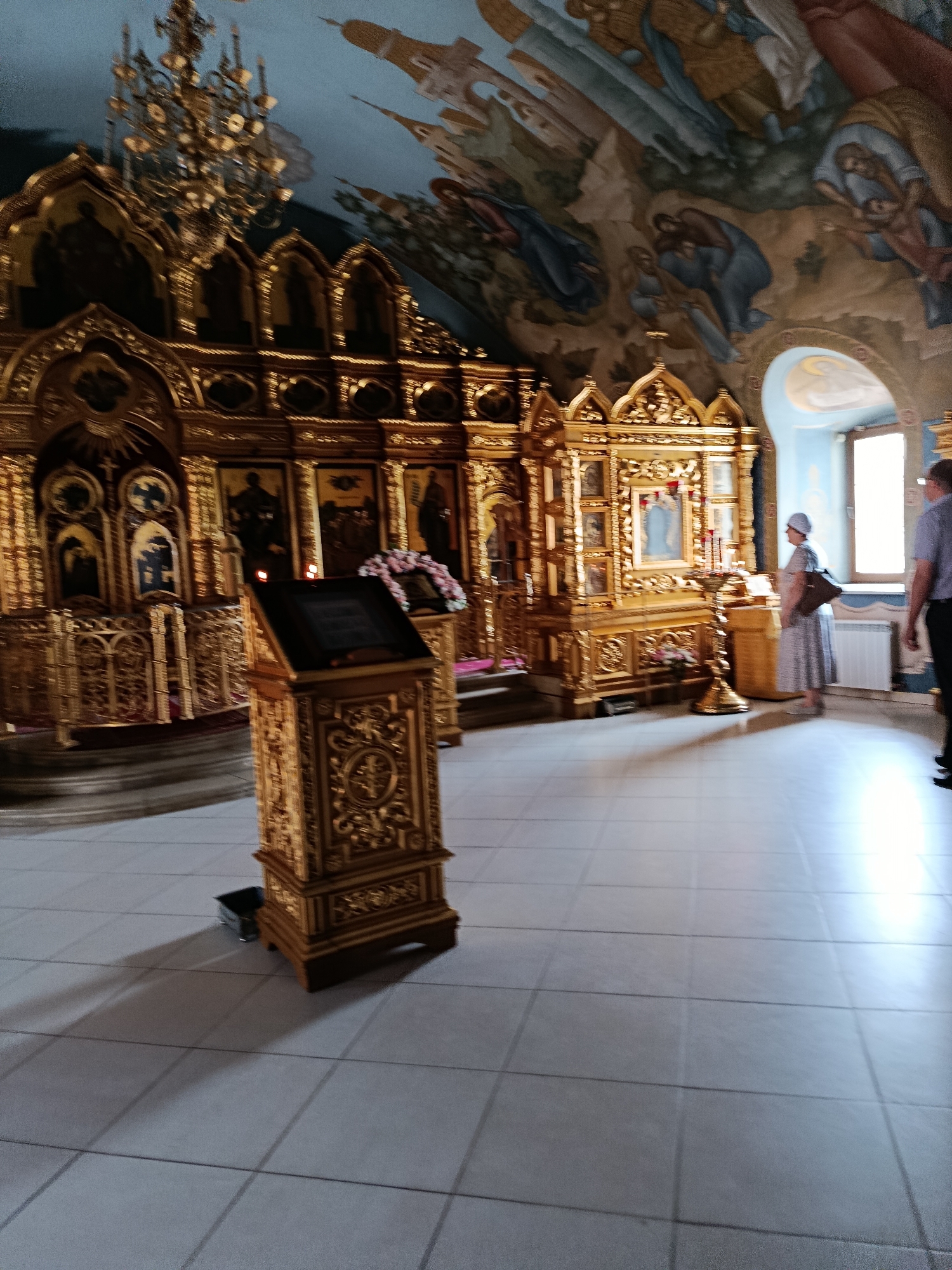 The image depicts the interior of a richly decorated Orthodox church, likely in an unknown location in Russia or a country with a similar architectural style. The scene is dominated by an ornate, gilded iconostasis, a wall of icons and religious imagery that separates the nave from the sanctuary. The iconostasis features intricate carvings and numerous icons, with a large central icon of the Virgin Mary. Above the iconostasis, the vaulted ceiling is adorned with colorful frescoes depicting biblical scenes and figures.
In the foreground, a wooden lectern with detailed carvings stands on a tiled floor. To the right, near a window, a woman in a light-colored headscarf and dress is standing, facing the iconostasis, with a brown bag on her shoulder. She appears to be in prayer or contemplation. Further to the right, a man in a light-colored short-sleeved shirt and dark trousers stands with his back to the viewer, also facing the iconostasis.
The floor is made of large, square tiles, reflecting the light from the window and the ornate decorations. A large chandelier hangs from the ceiling, adding to the opulence of the space. The overall impression is one of solemnity and grandeur, typical of Orthodox church interiors.